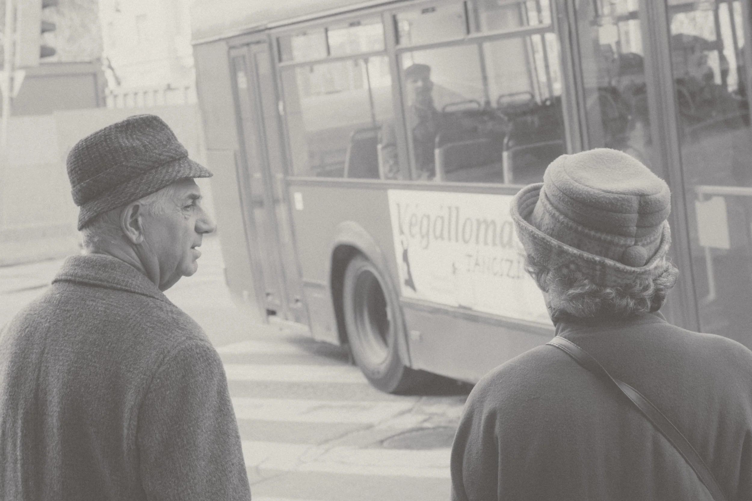 Two elderly people wearing hats standing on a city sidewalk near a bus with an advertisement.