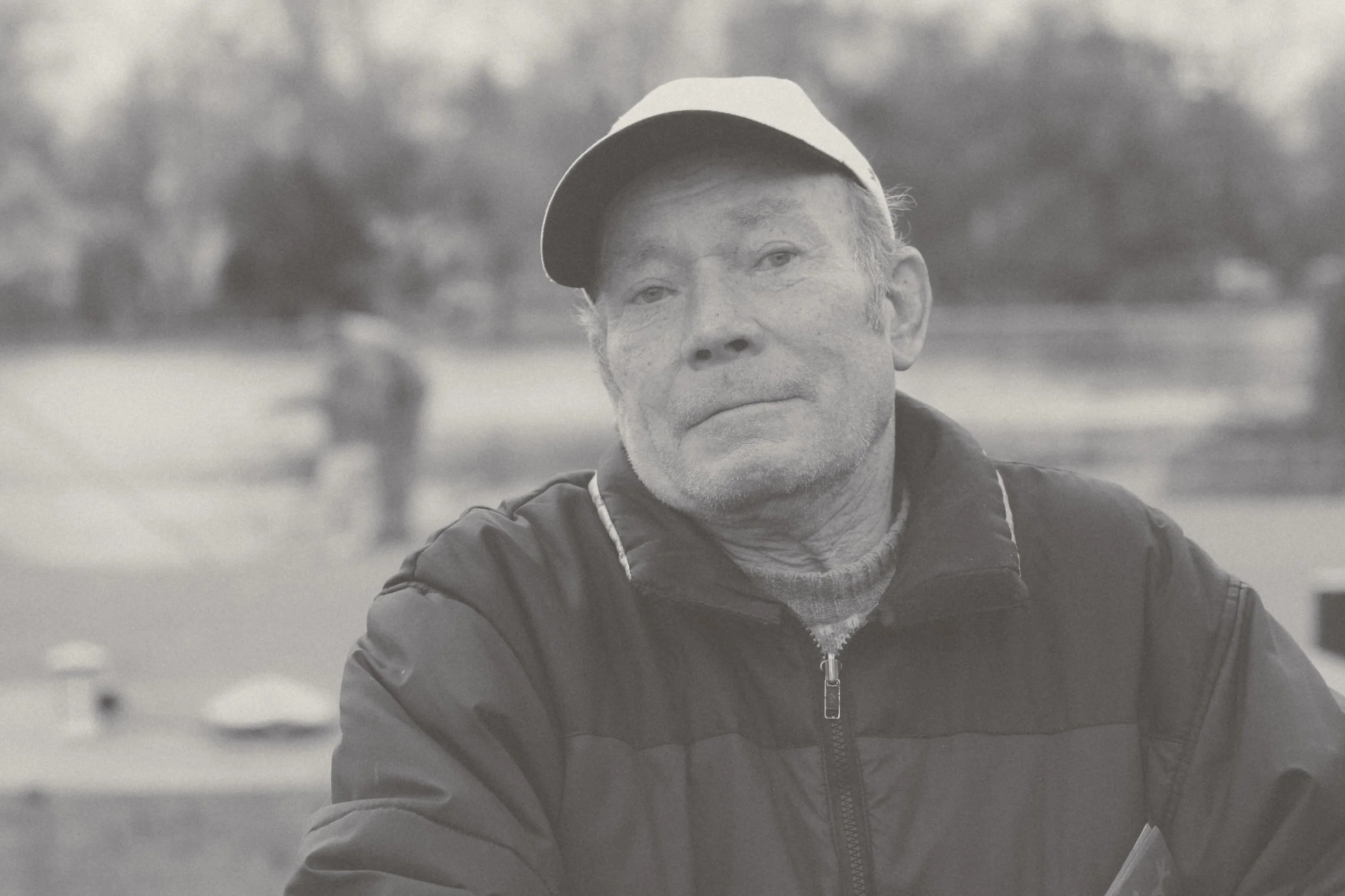 A black and white photo of a man wearing a cap and a zipped jacket outdoors, with blurred background trees and a person in the distance.