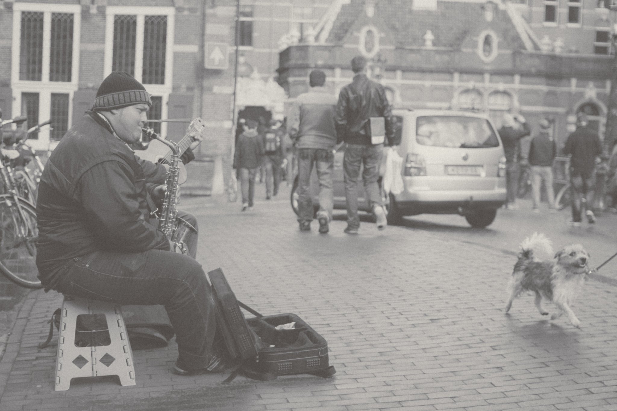 A man playing the saxophone on a sidewalk in an urban area, with people walking and a dog on a leash nearby, and a car parked behind them.