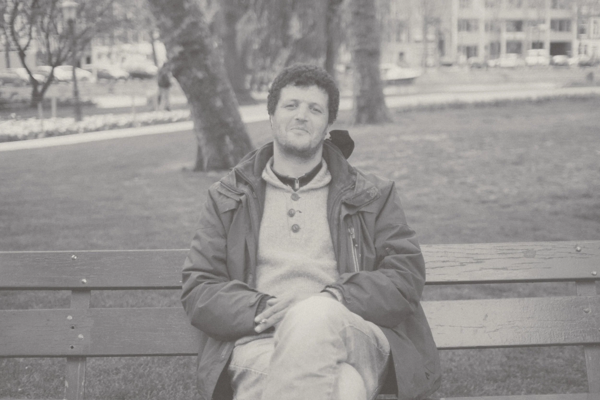 A man sitting on a park bench in an outdoor setting with trees and buildings in the background