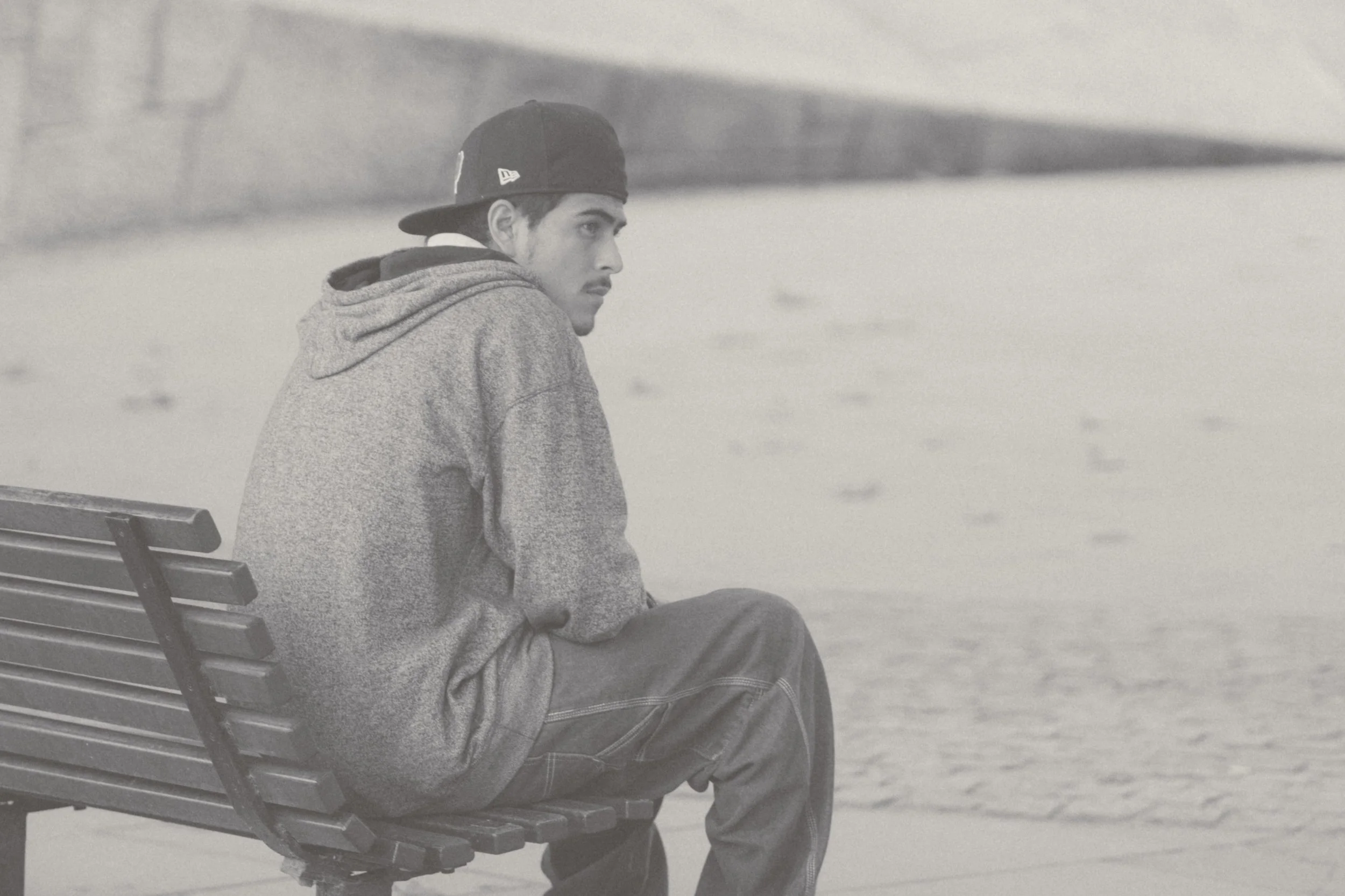 A young man with dark hair, wearing a backward baseball cap and gray hoodie, sitting alone on a park bench beside a body of water, looking pensively to the side.