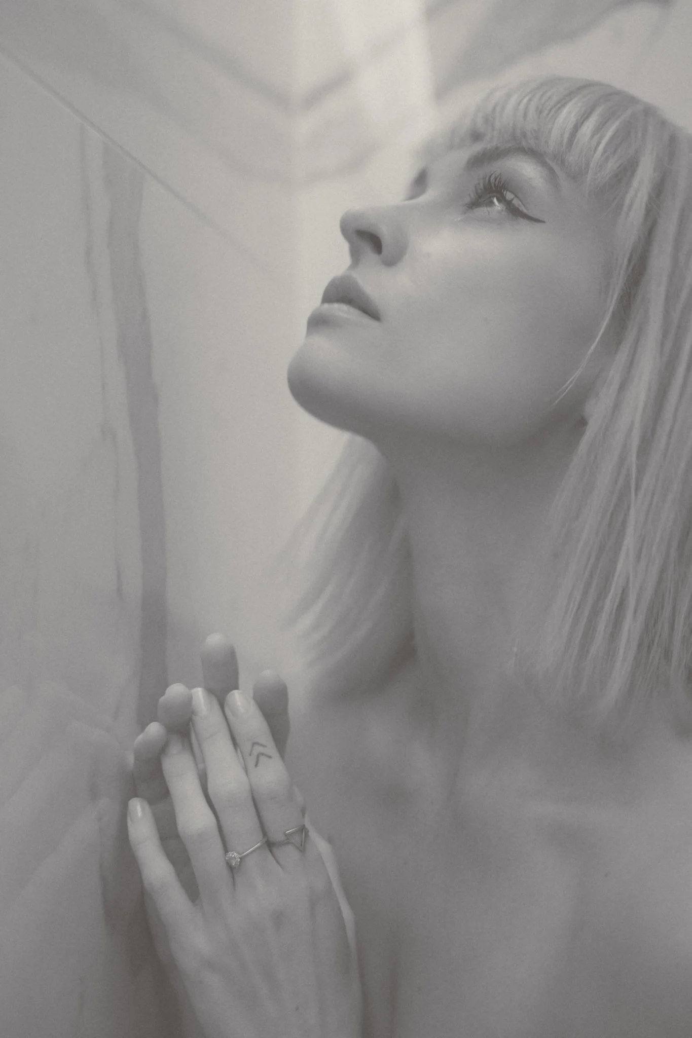 Black and white photo of a mannequin's head with styled hair, and hands with rings, holding some objects close to its face, in a reflective surface.