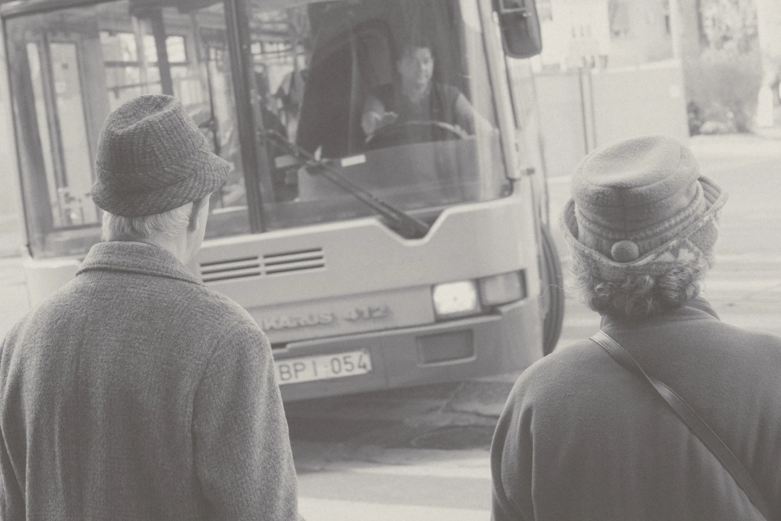 Two elderly women with hats waiting at a bus stop as a bus approaches.