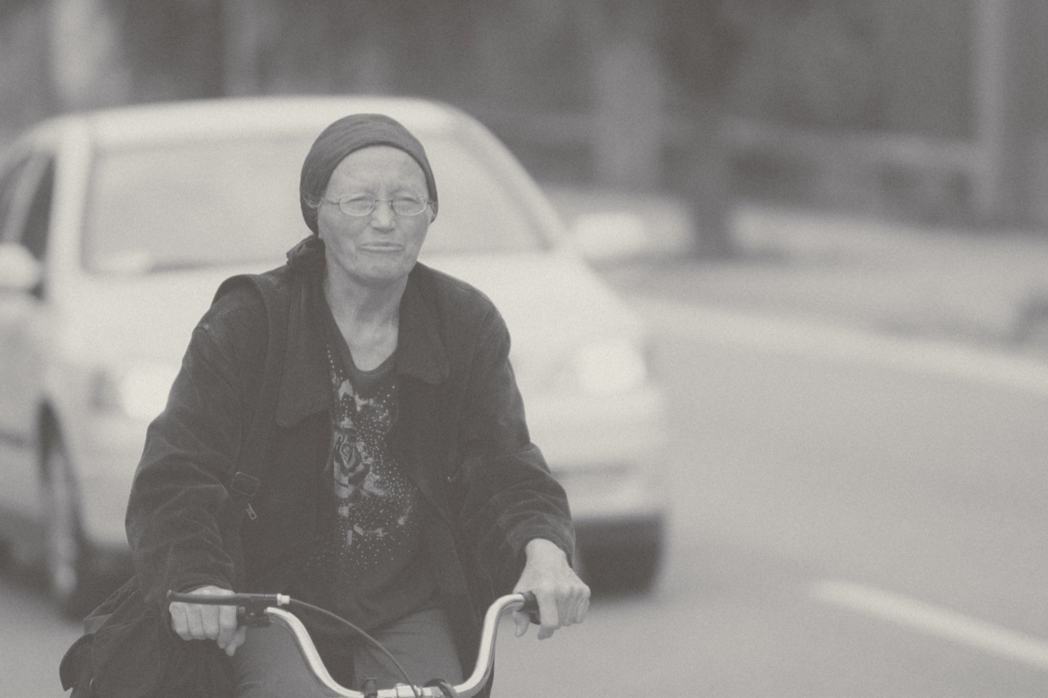 An elderly person with glasses and a beanie riding a bicycle on a city street, with cars blurred in the background.