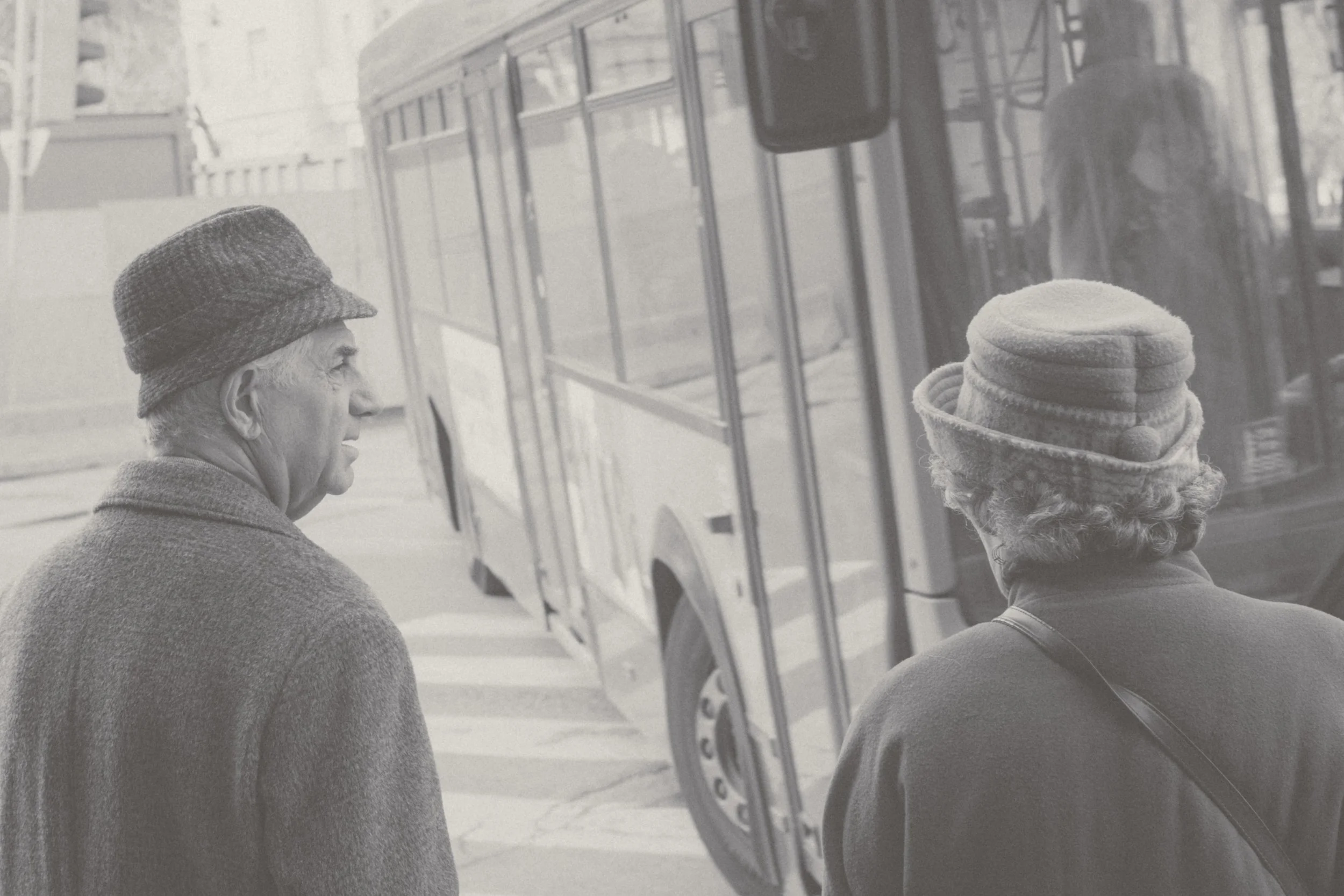 Two elderly women with curly hair, wearing hats and coats, standing on the sidewalk near a bus, engaging in conversation.