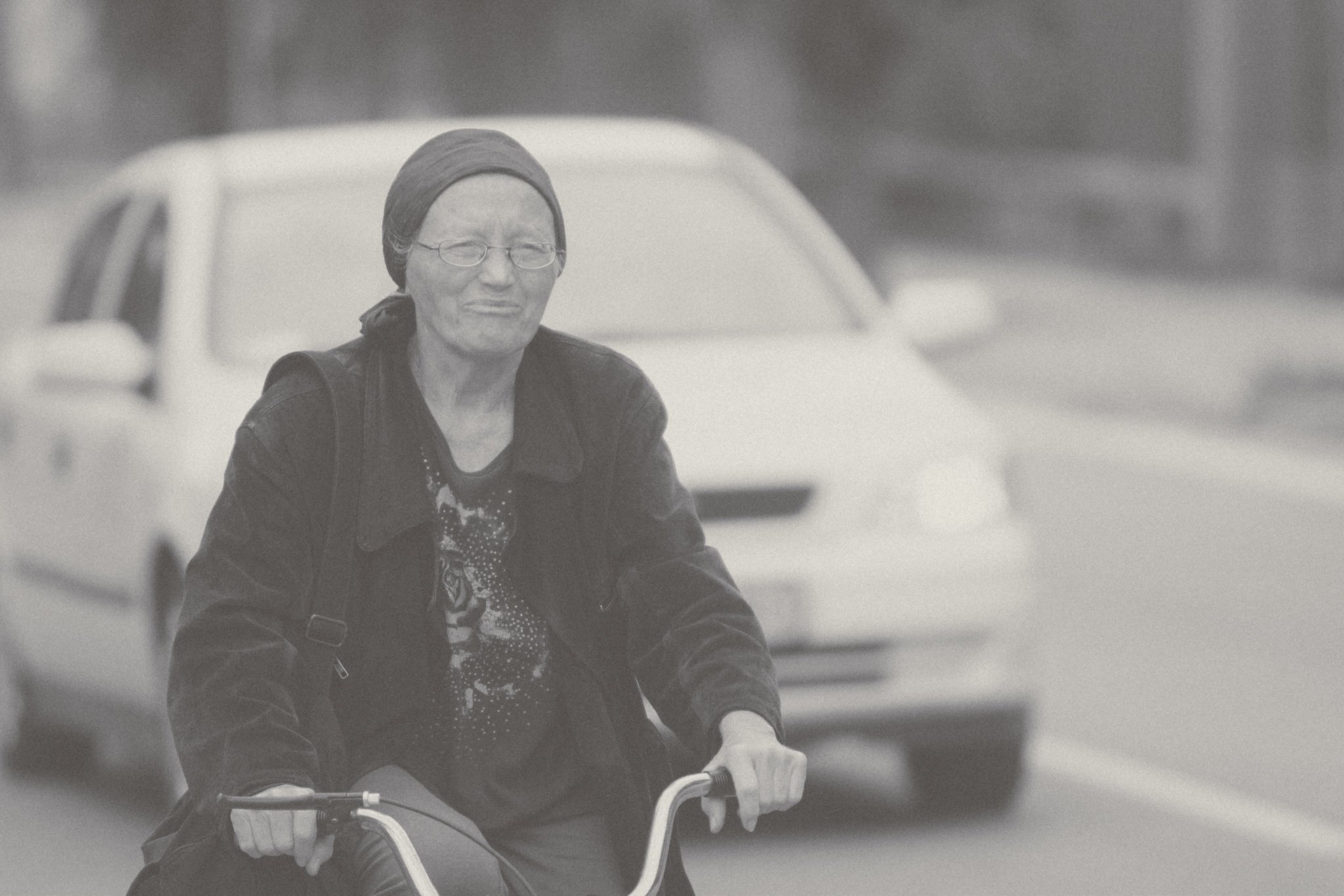 An older woman with glasses, wearing a dark jacket and headscarf, riding a bicycle on a street with parked cars in the background.
