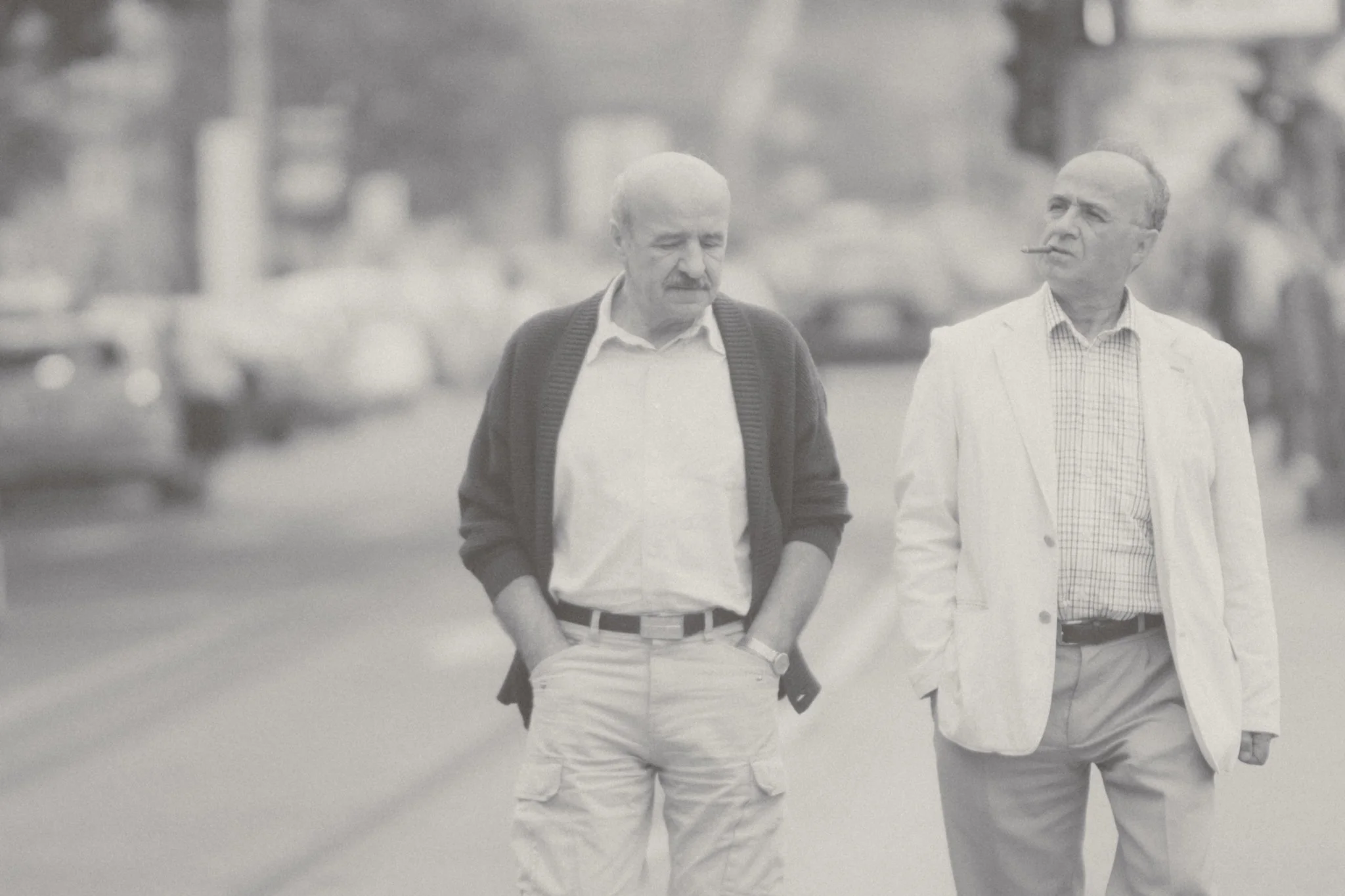 Two older men walking on a street, one smoking a cigarette, dressed in casual and light-colored clothing, with cars and blurred background.