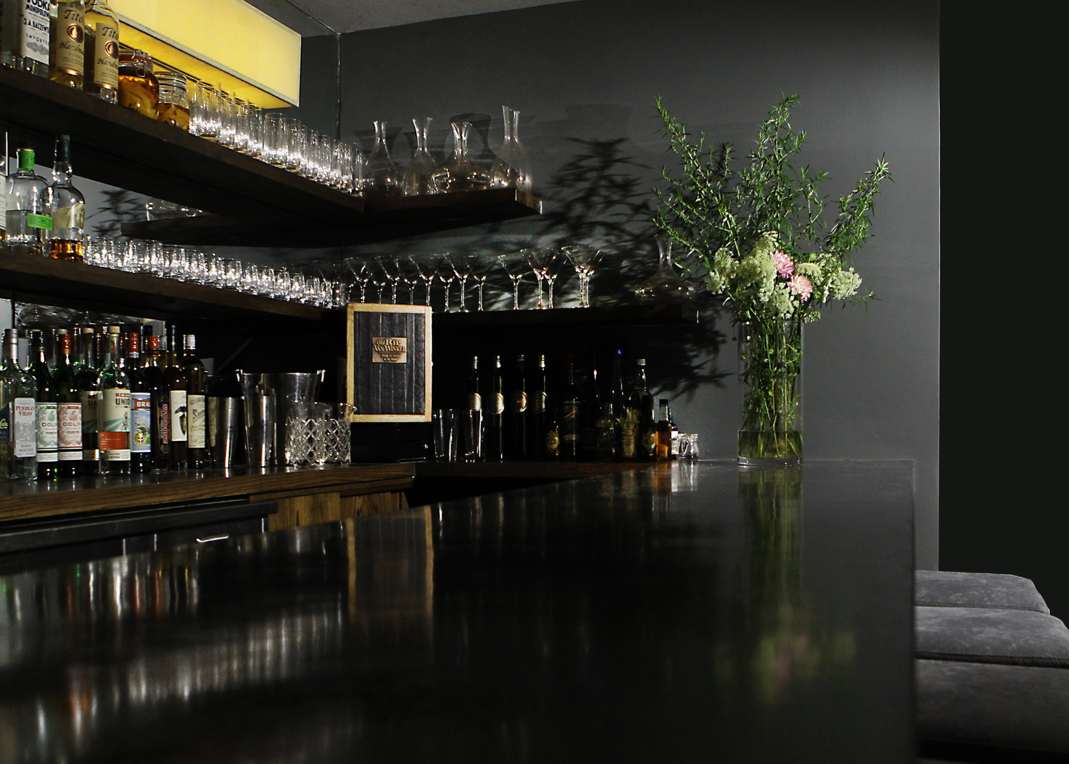 The front bar features a new custom poured black concrete bar top, with wrap-around floating oak shelving behind and John Wigmore custom light-fittings on a mirrored backdrop.