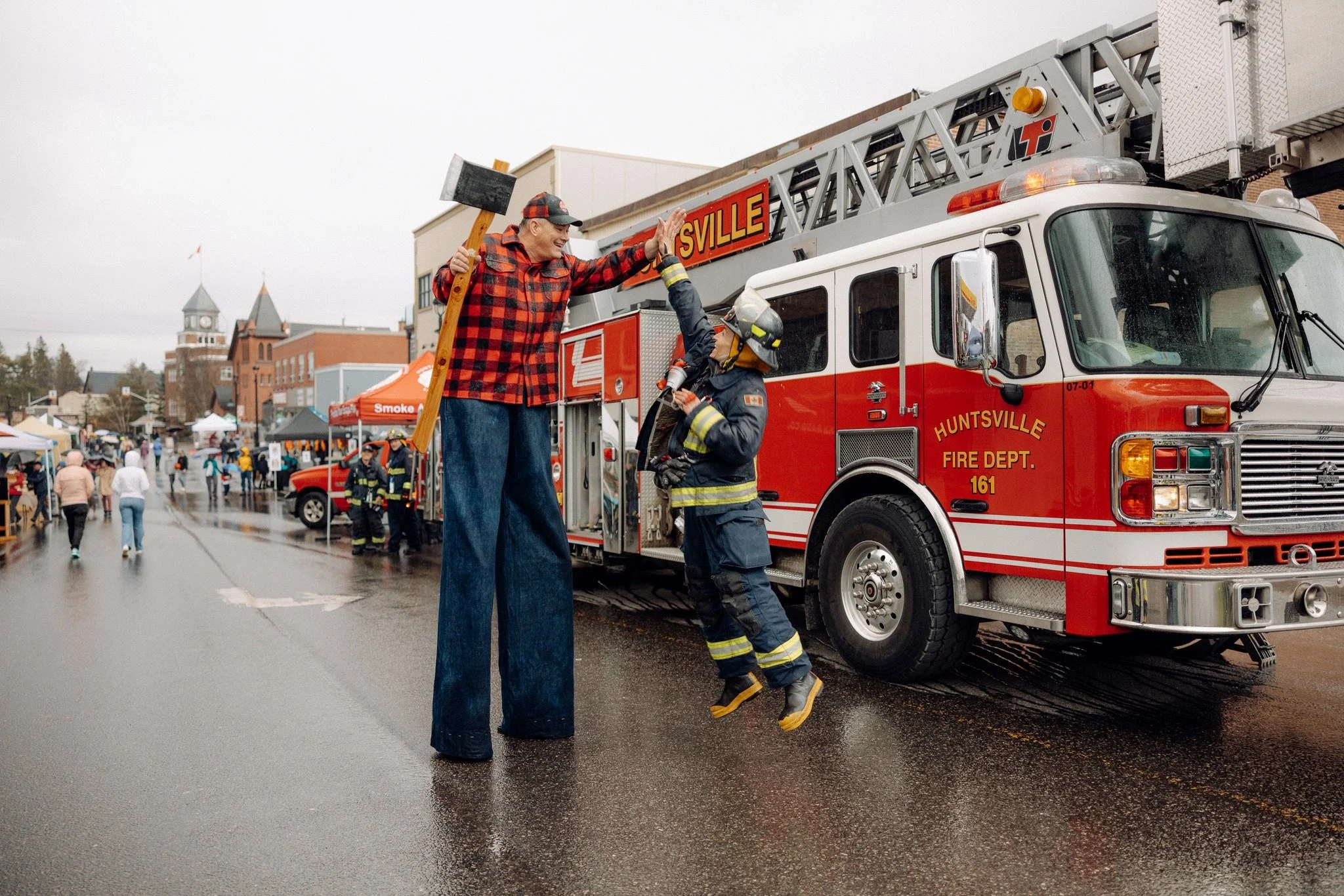 A person on tall stilts dressed as a firefighter high-fiving a young firefighter in uniform next to a red Hunterville fire truck during a rainy day event in town.