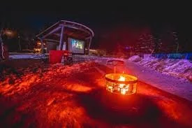 Snowy outdoor scene at night with a fire pit and ice-covered ground, illuminated by colorful lights