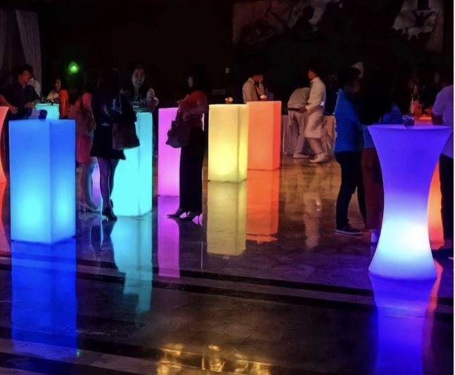 People gather around illuminated, color-changing cocktail tables in a dark venue.