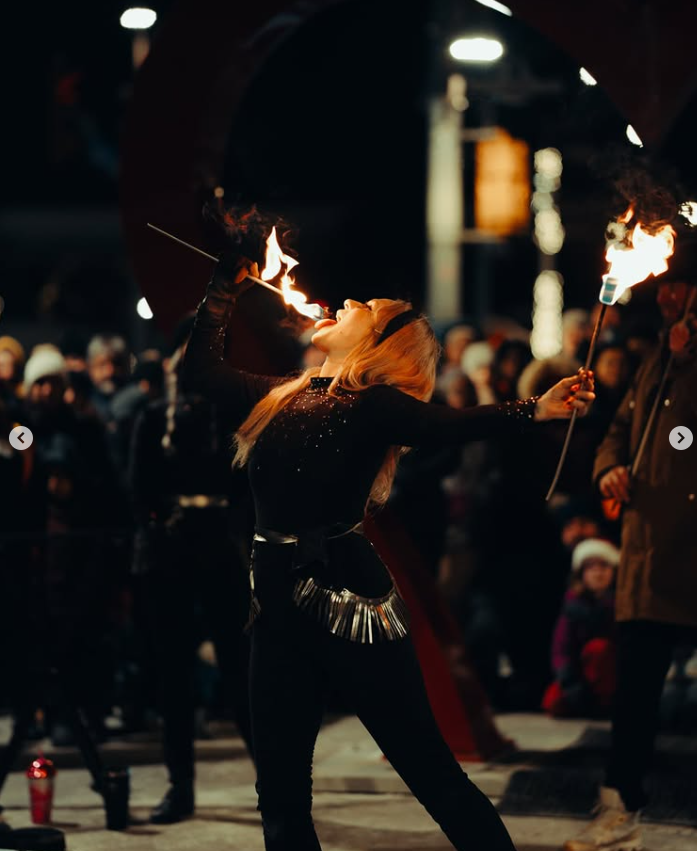 A fire performer juggles flaming torches in front of a crowd at night, with a large orange sculpture or structure behind them.