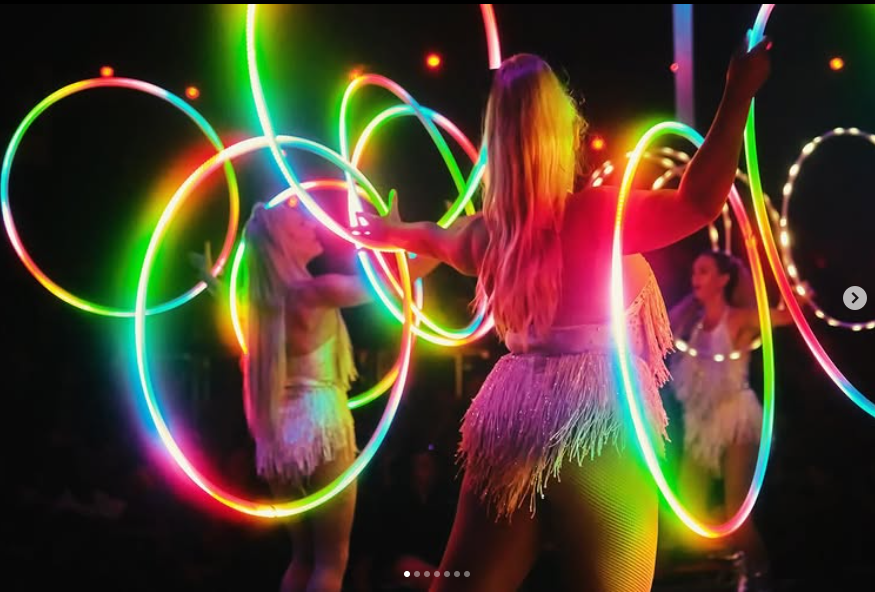 Women dancing with colorful glowing hula hoops in a dark indoor setting, with vibrant neon lights creating a lively atmosphere.