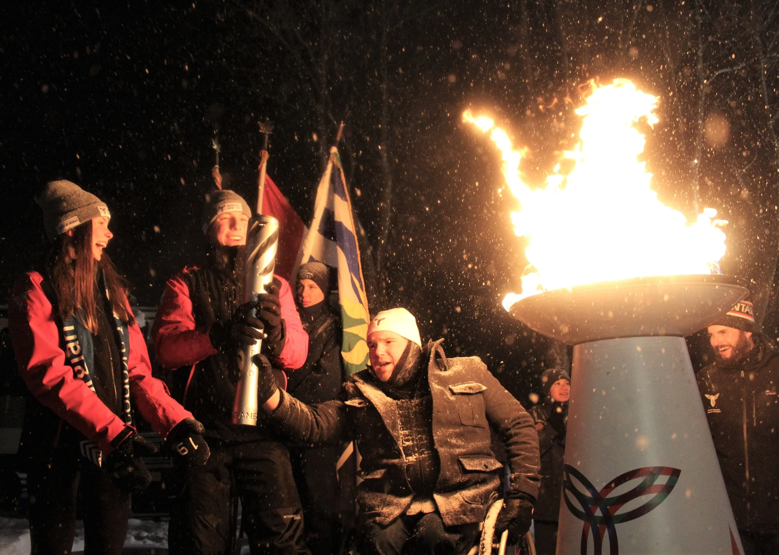 Group of people gathered around a torch with flames at night, celebrating, with flags in the background.