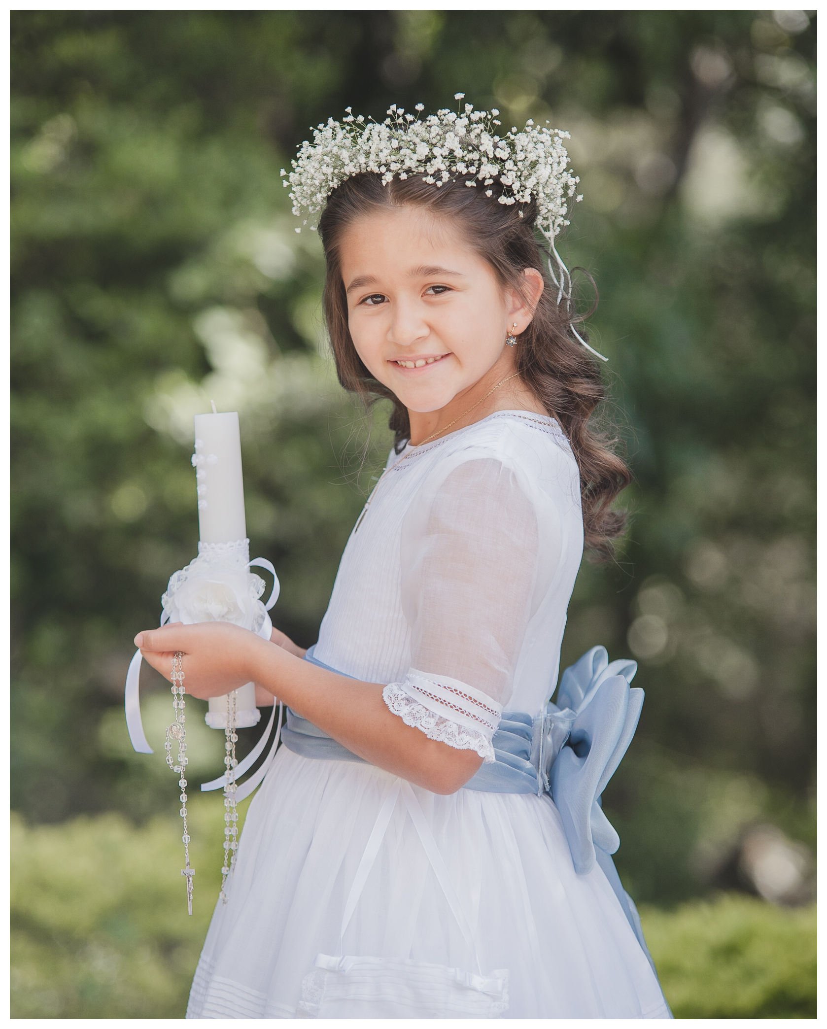 Retrato de una niña sonriendo con su vestido de primera comunión, sosteniendo una vela decorada y un rosario en sus manos.