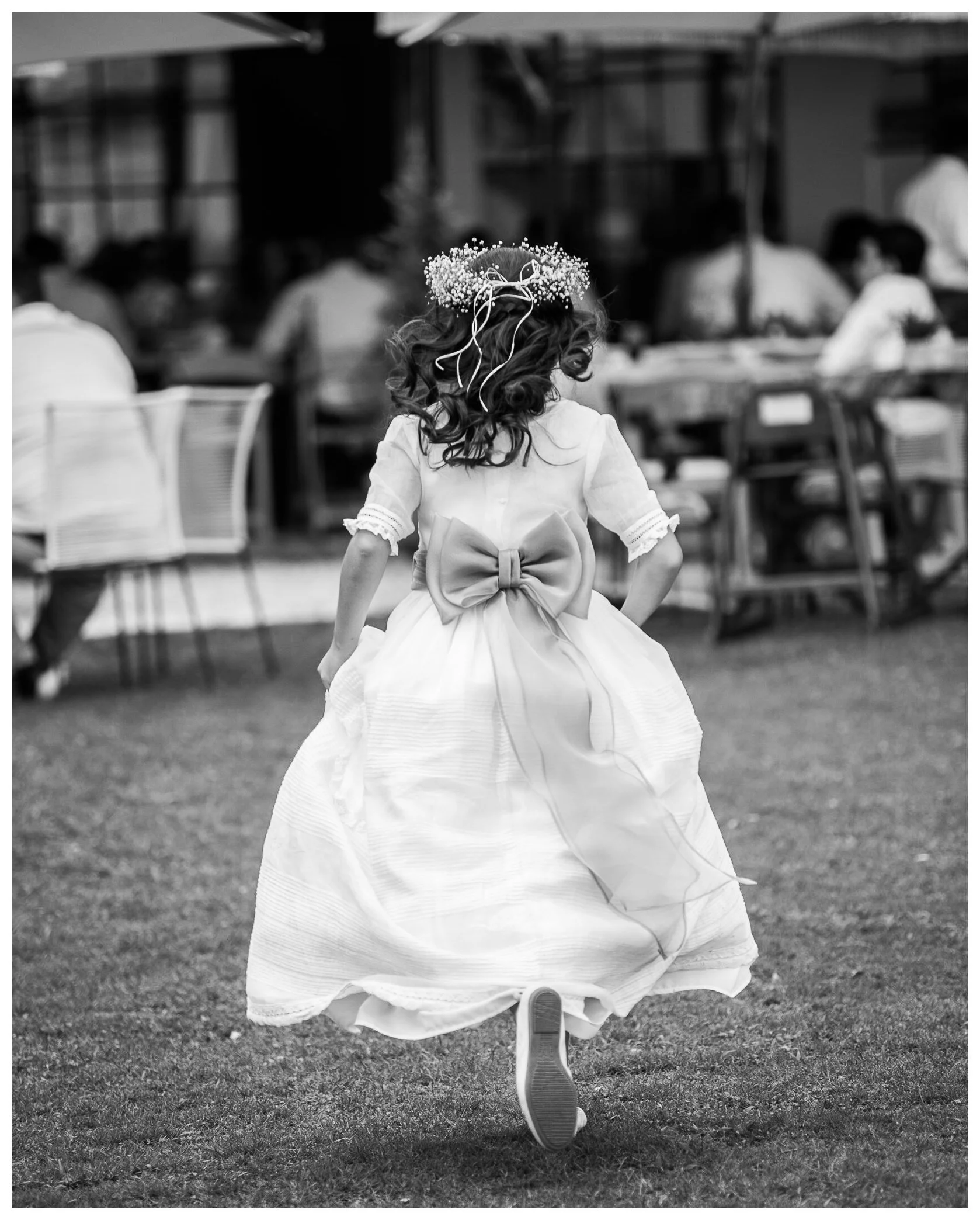 Fotografía en blanco y negro de una niña con su vestido de primera comunión corriendo por un jardín, vista desde atrás.