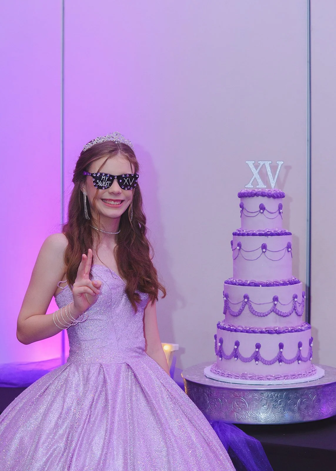 Quinceañera en Guatemala posando divertida junto a un pastel de 15 años morado de cuatro pisos, usando lentes festivos de XV años.