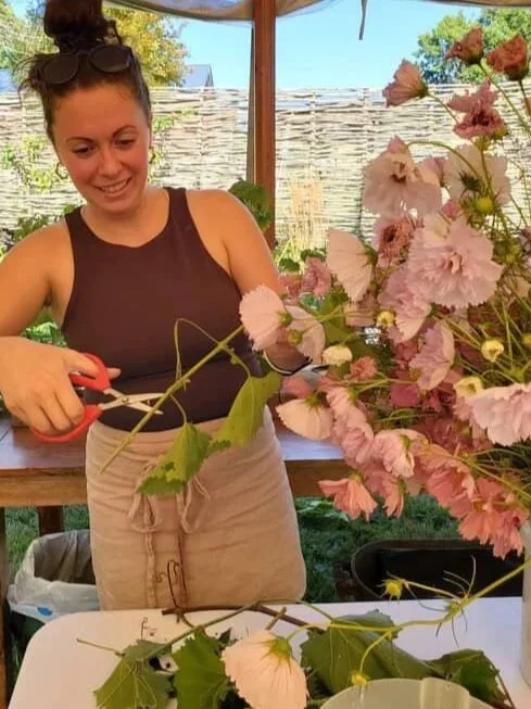 A woman wearing an amphitrite linen apron smiling while trimming light pink and purple flowers in a garden or greenhouse setting.