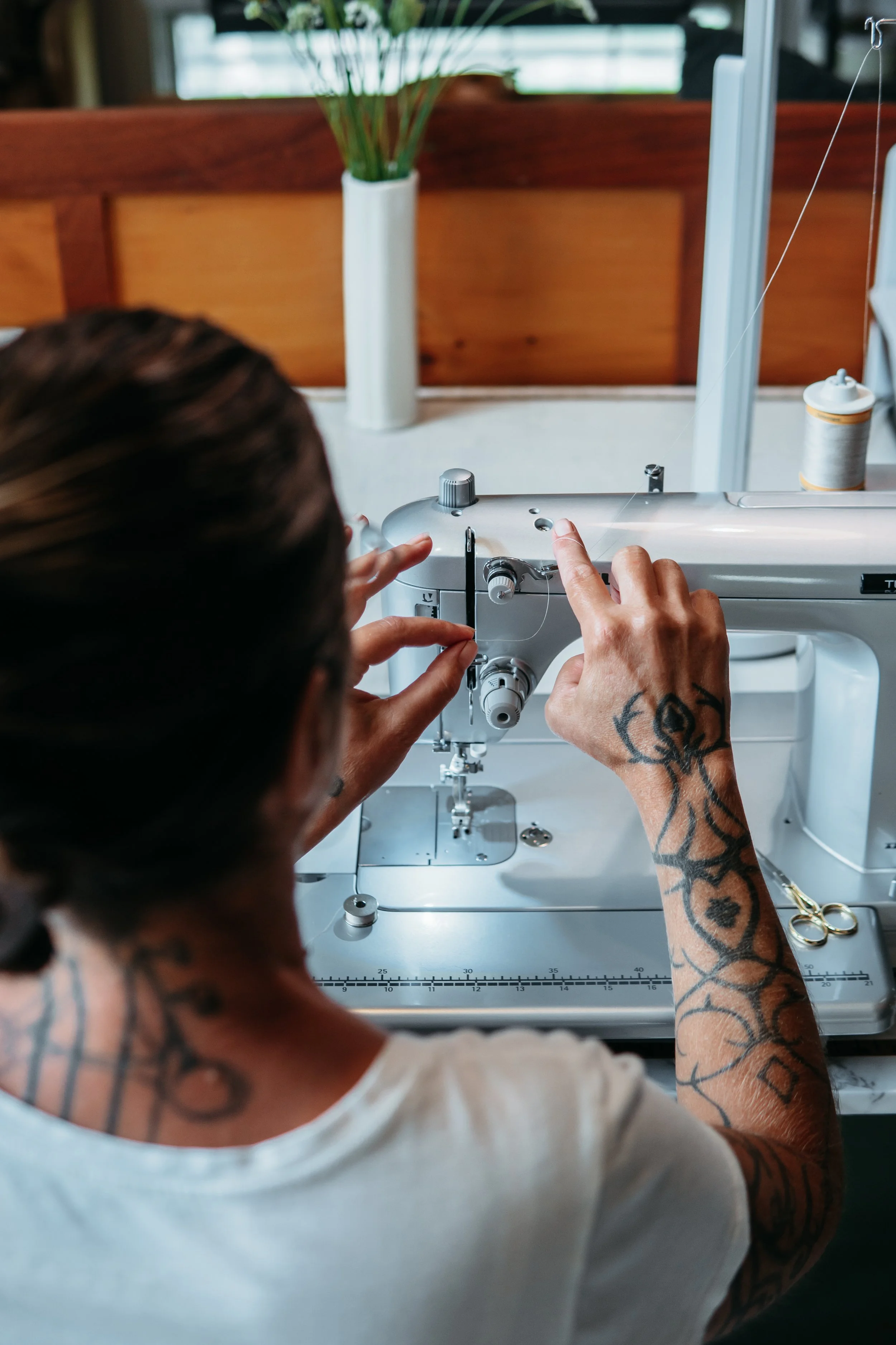 Katrina Kelley, Seamstress and owner of amphitrite studio linens... A person with tattoos is sewing on a Juki sewing machine. In the background, there is a wooden panel and a white vase with green plants on a windowsill.