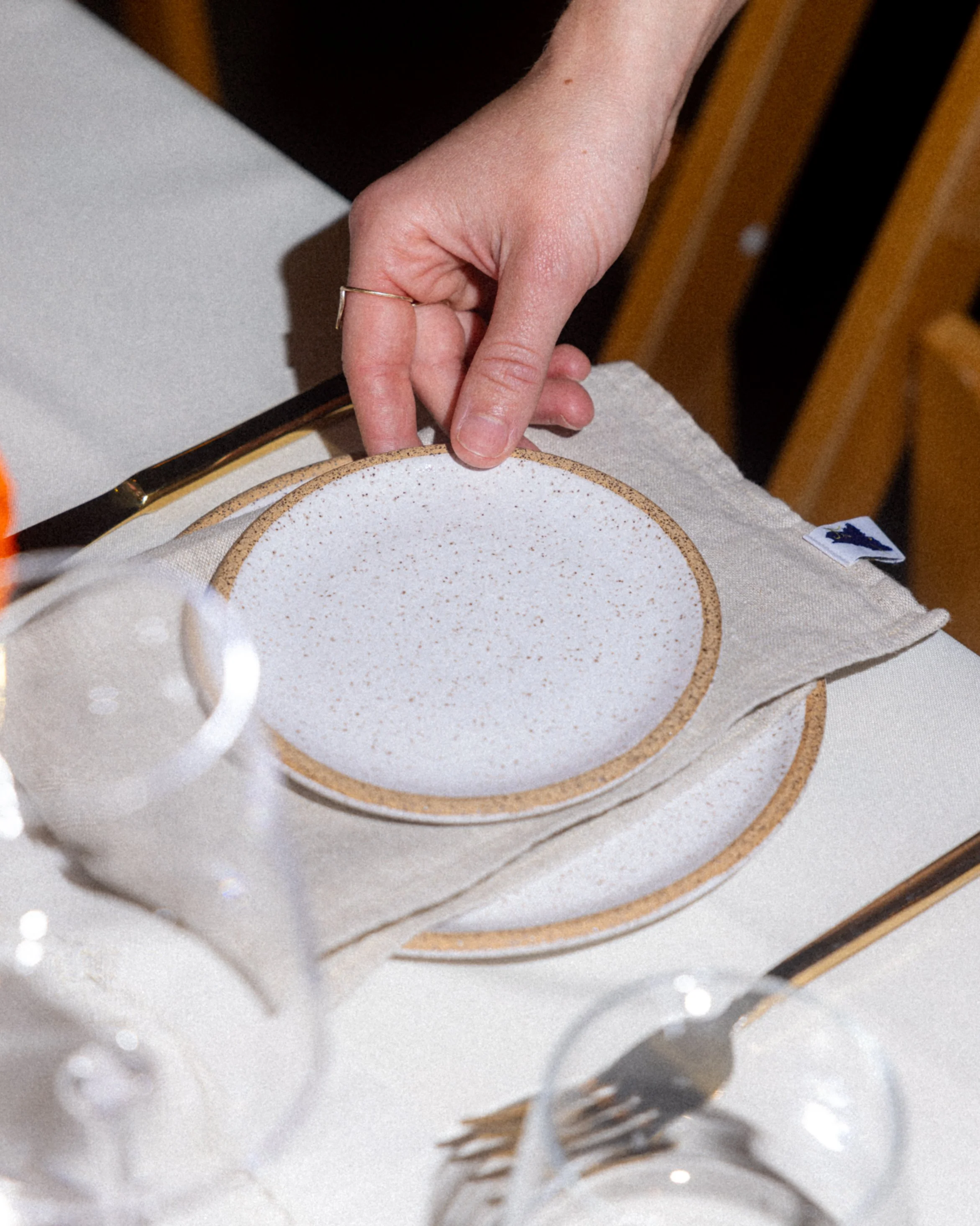 Hand placing a small ceramic plate on a beige napkin, with table setting including a fork and glass.