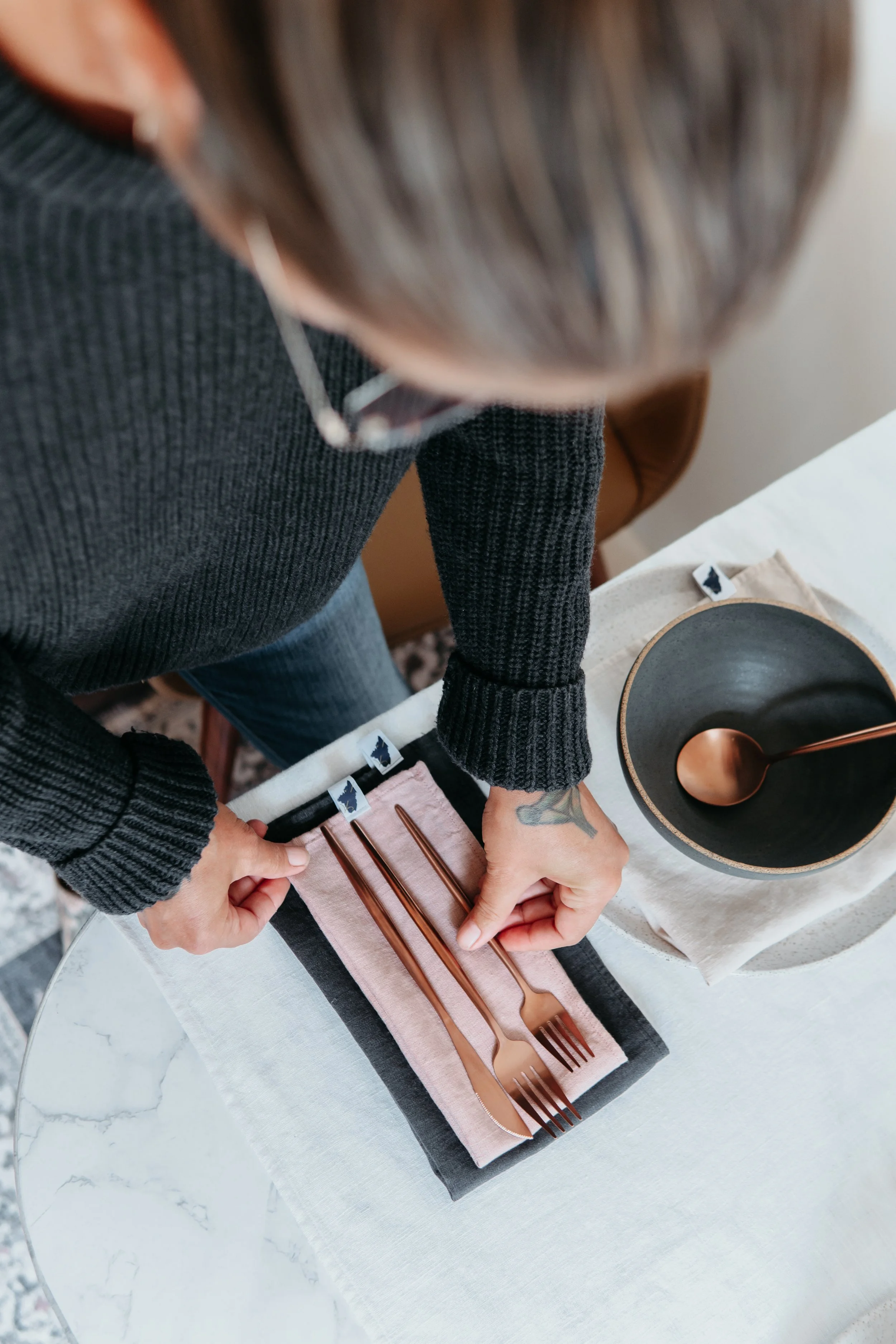 Person arranging copper-colored forks on a rose linen amphitrite cloth napkin on a white marble table.