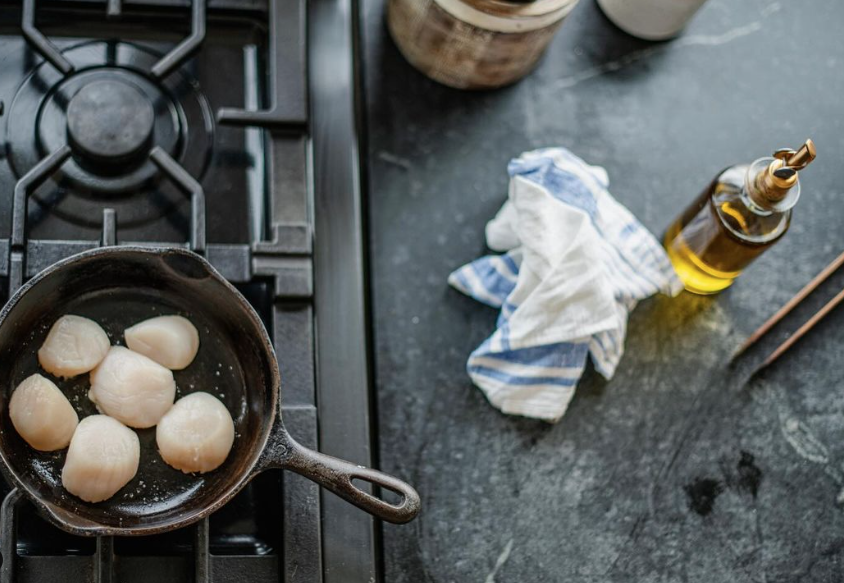Raw scallops in a cast iron skillet on a stovetop, olive oil bottle, and striped towel on counter.