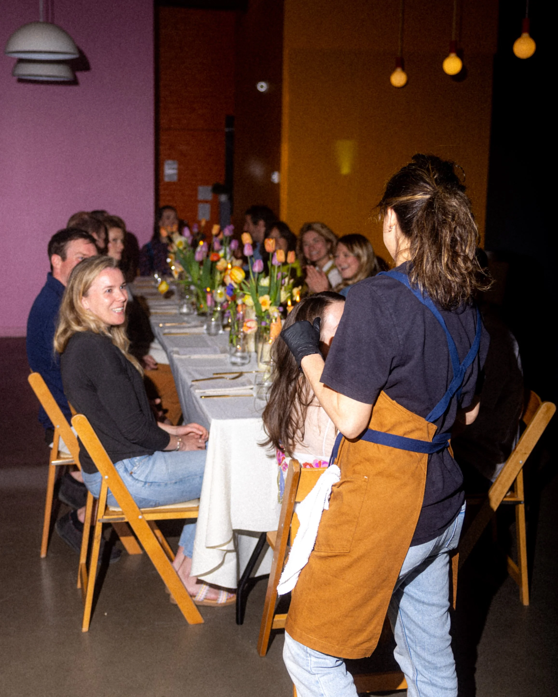 Group of people seated at a long dining table with flowers, served by a person wearing an apron.