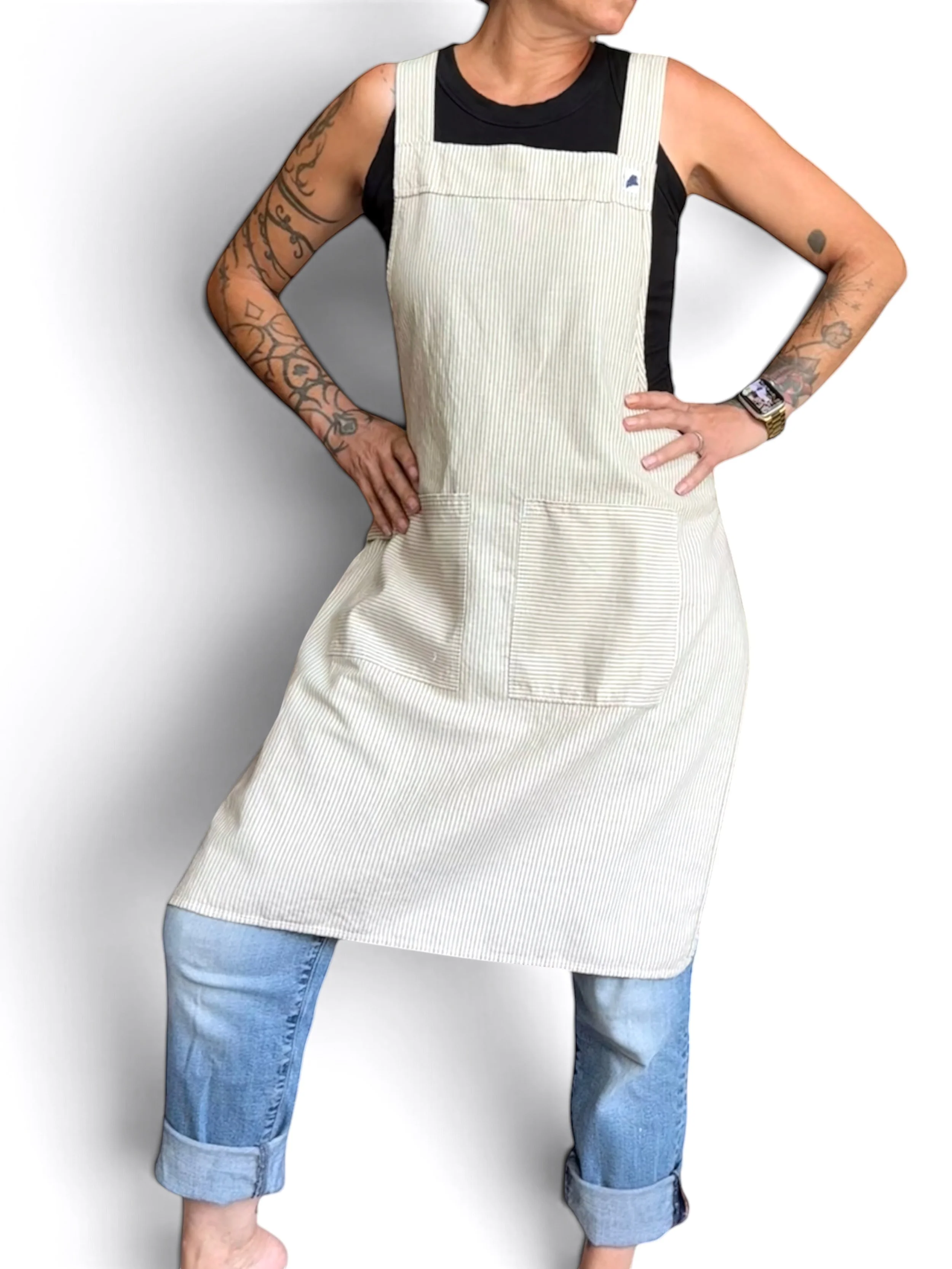 A person with tattoos on arms wearing a black sleeveless shirt, a light-colored pinstriped amphitrite apron, and rolled-up blue jeans standing against a white background.