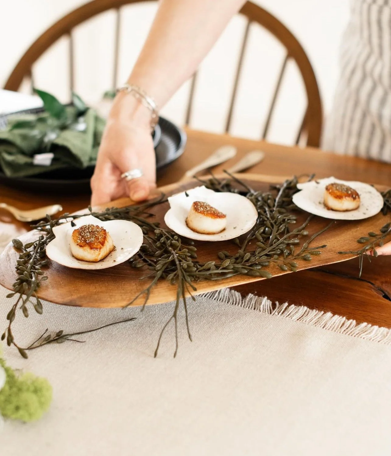 Hand holding a tray with scallops in shells, garnished with herbs, on a table with cutlery and a cloth.