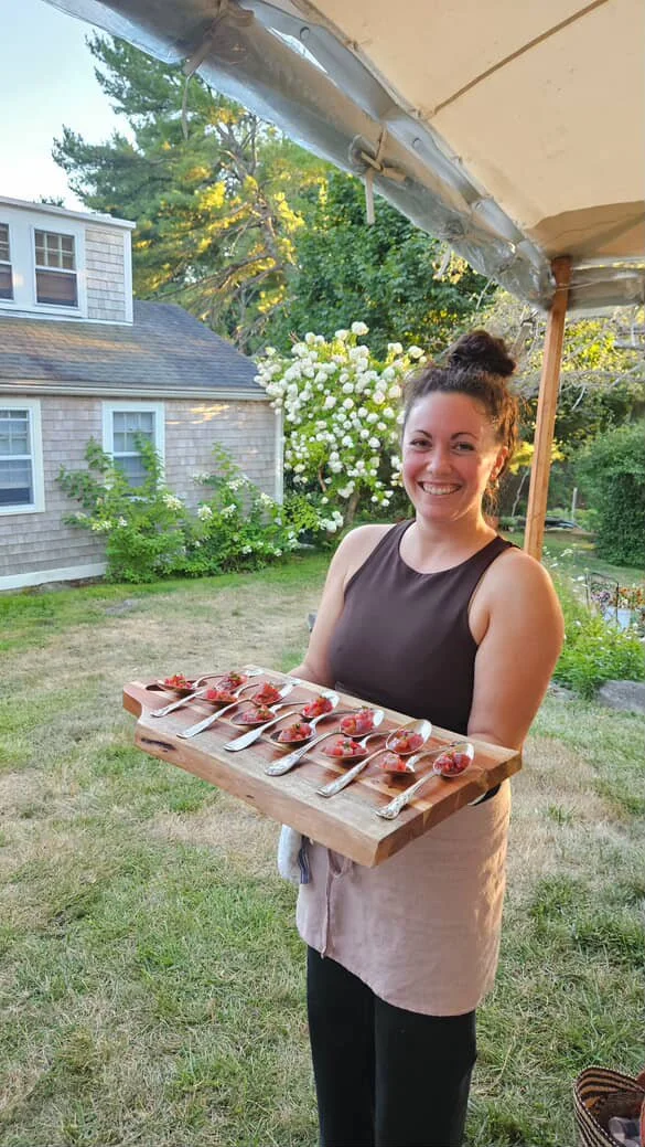 A woman wearing an amphitrite linen apron, holding a wooden serving board with ten spoons, each filled with a small serving of hors d'oeuvres, standing outdoors under a white patio umbrella with a garden and house in the background.