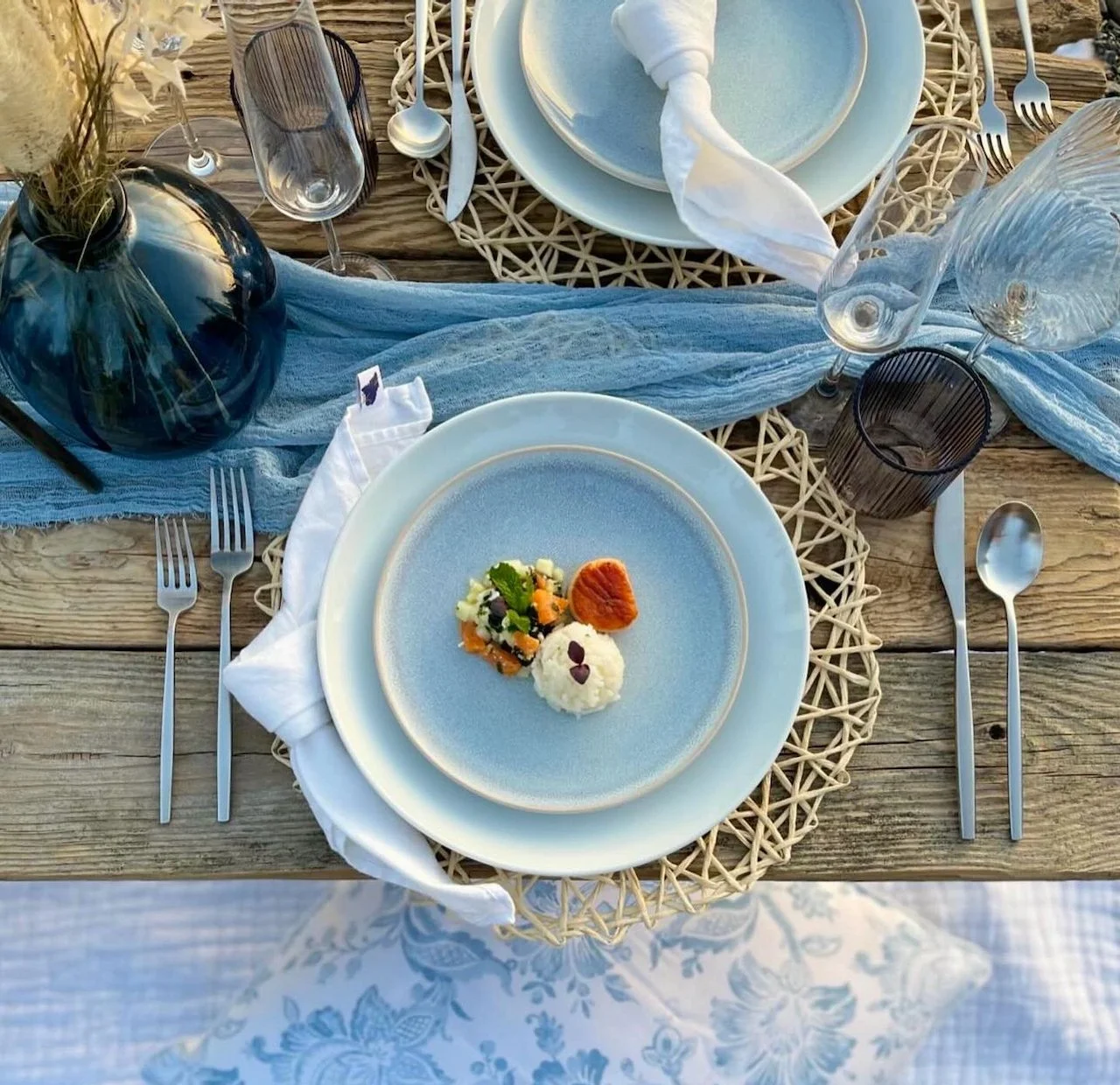 Elegant table setting with blue plates, white napkins, and gourmet dish on a rustic wooden table. Decor includes a glass vase with flowers, blue fabric runner, and assorted glassware and cutlery.