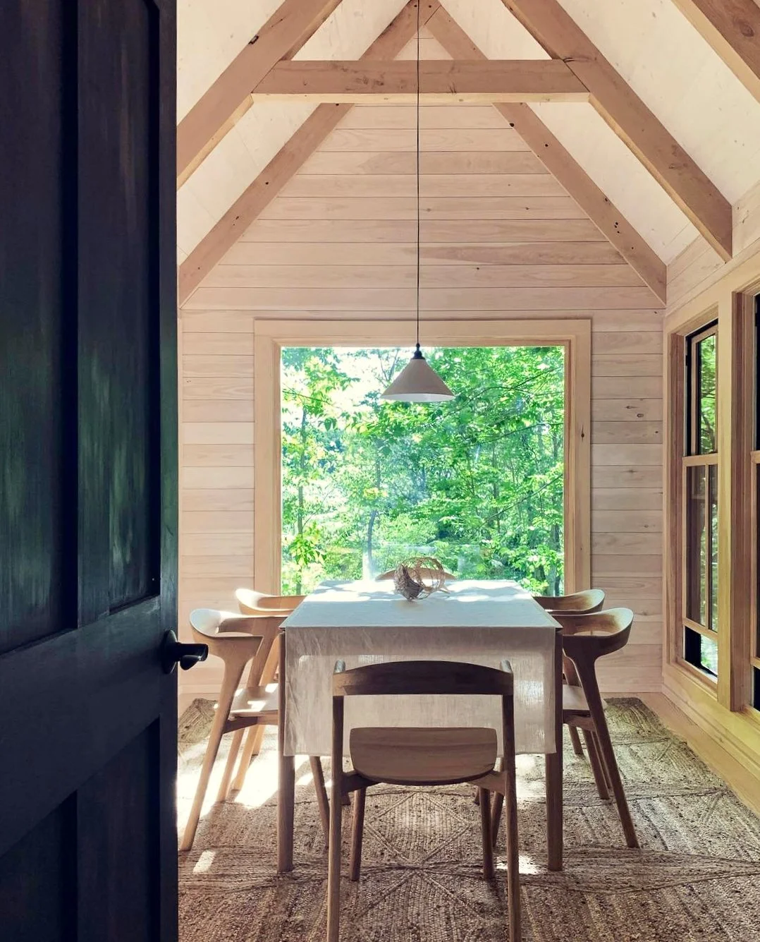 Inside a wooden cabin dining area with a view of lush green trees through a large window, featuring a rectangular table with a white amphitrite tablecloth, surrounded by four matching wooden chairs, and a single hanging light fixture.