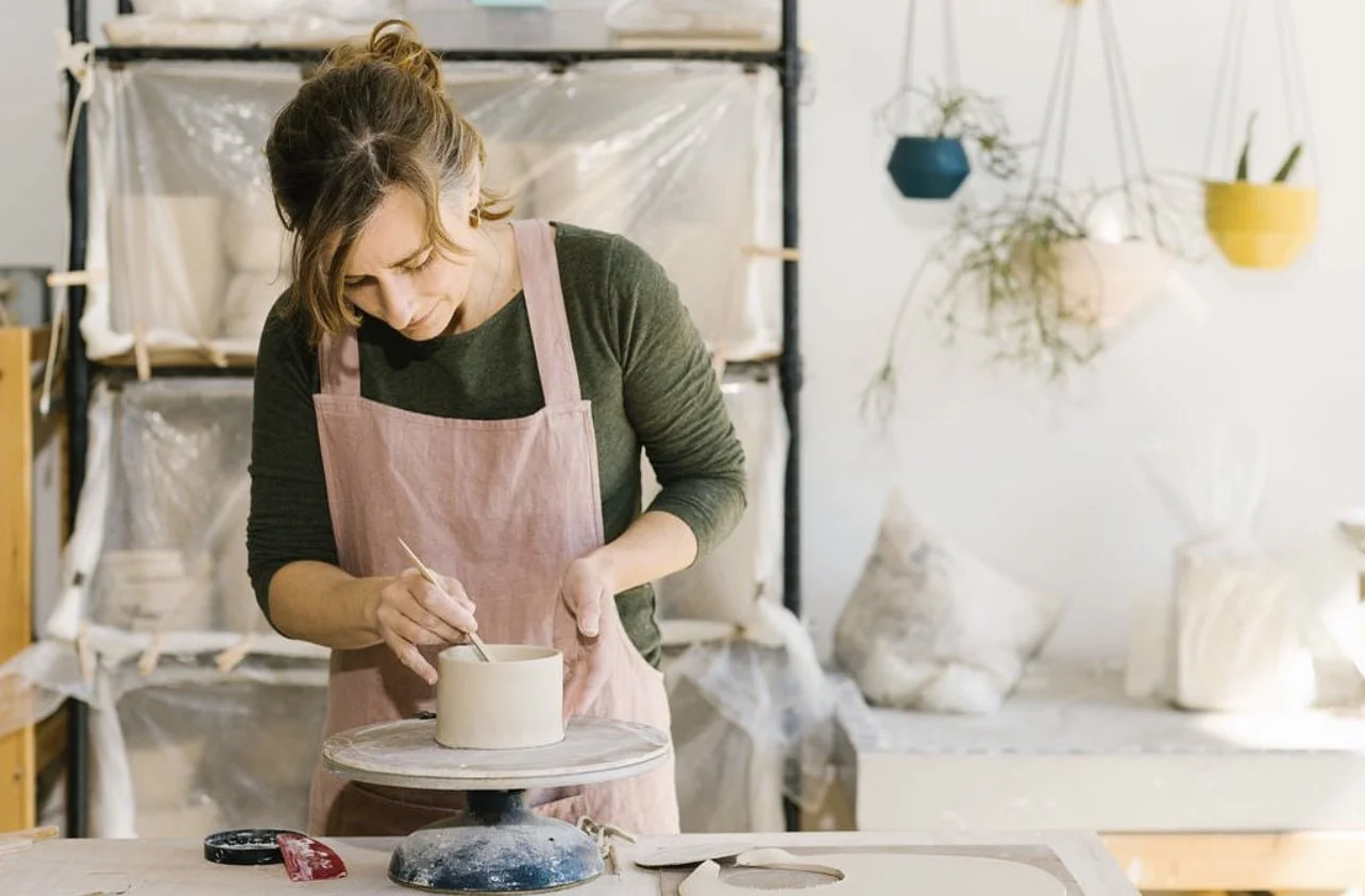 Woman shaping a ceramic pot in a studio workspace,  Linen apron shown.