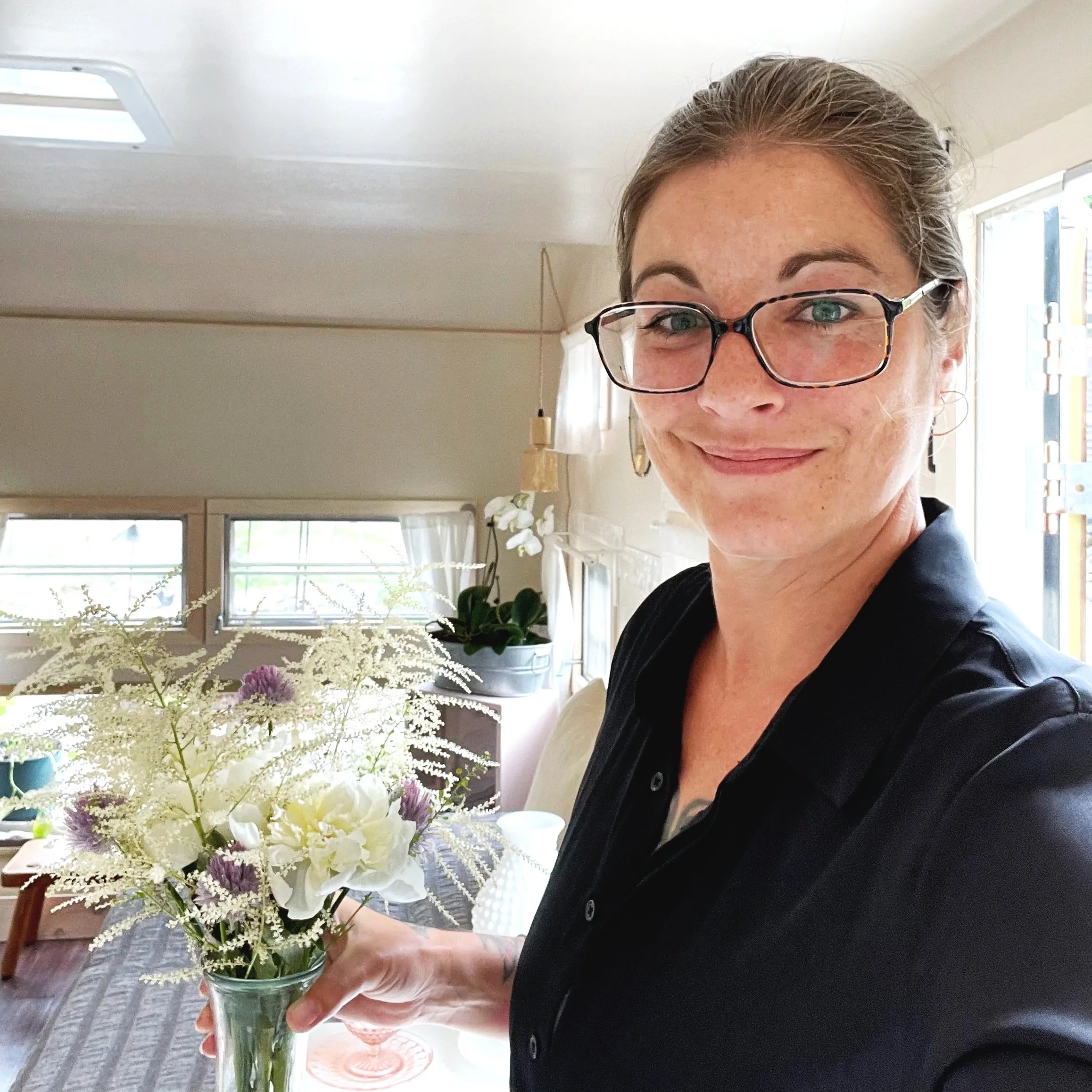 Katrina Kelley, Seamstress & owner of amphitrite linens studio, wearing glasses, smiling, holding a glass jar with a bouquet of flowers, including white and purple blooms, in a bright room with large windows.