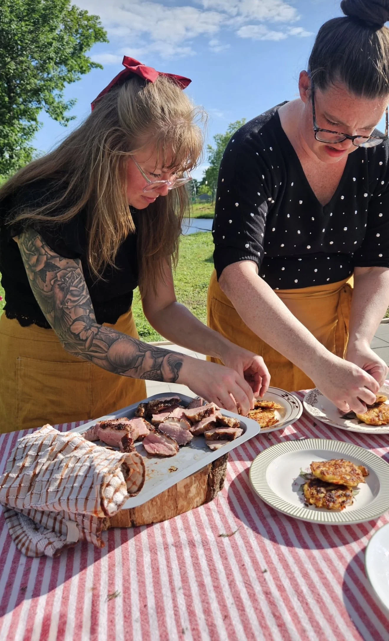Two women with glasses and tattoos are preparing food at an outdoor picnic table with a red and white striped tablecloth, with a scenic lakeside background, carving and serving cooked meat and pizza slices.  Linen apron shown.