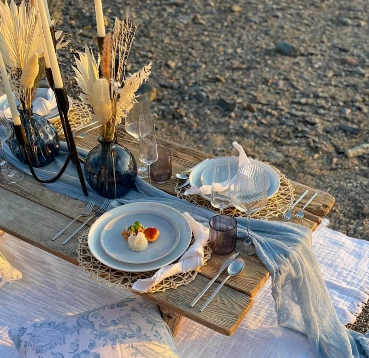 Outdoor rustic table setting with wooden bench, blue vases with dried flowers, plates, glasses, and a blue cloth runner.