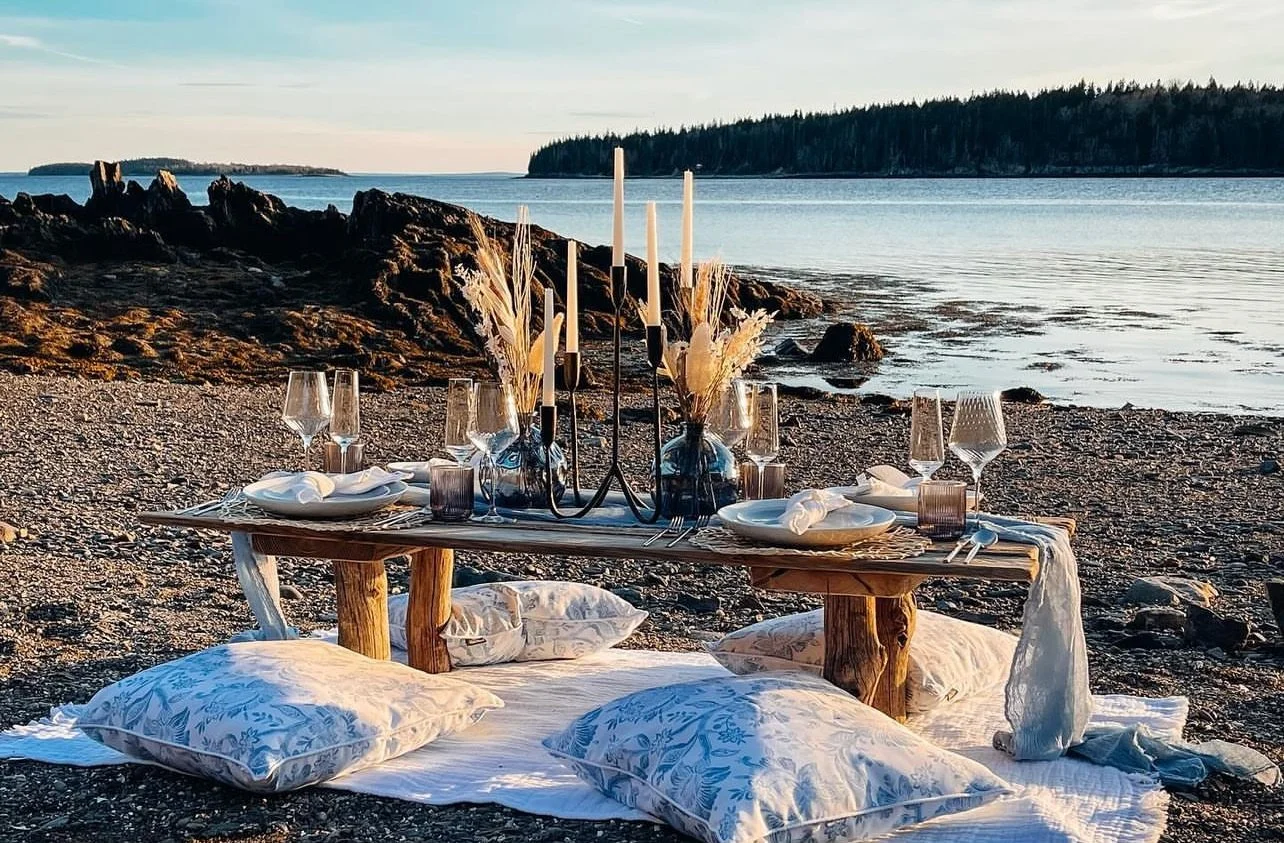 Beach picnic setup with a low wooden table, elegant tableware, candles, and decorative vases. Surrounded by cushions, set against a backdrop of rocky shoreline and water.