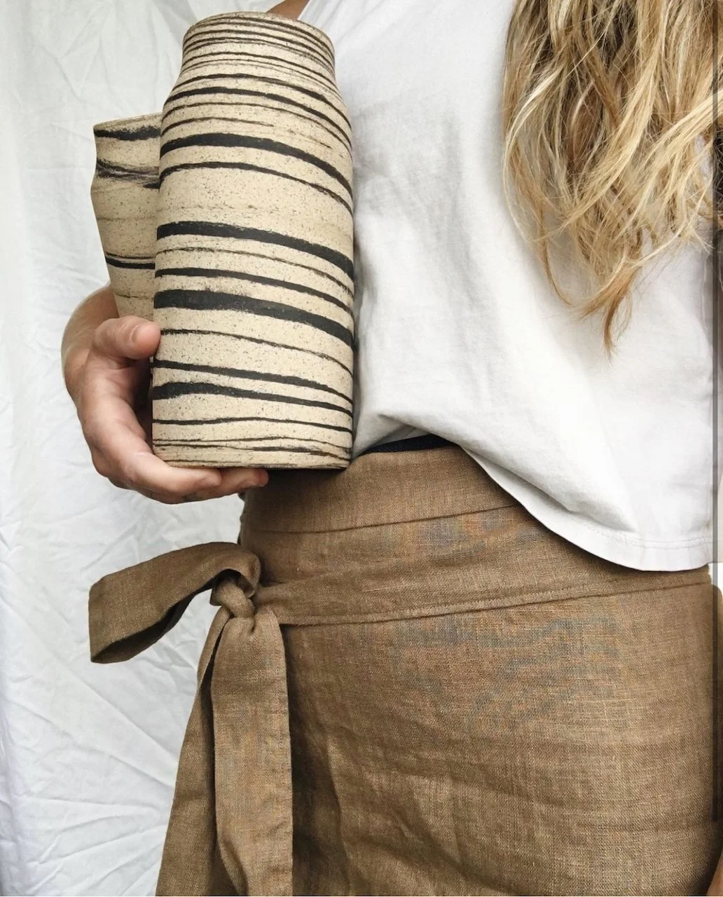 Person holding a stack of beige cups with black horizontal stripe patterns, wearing a white top and brown apron.