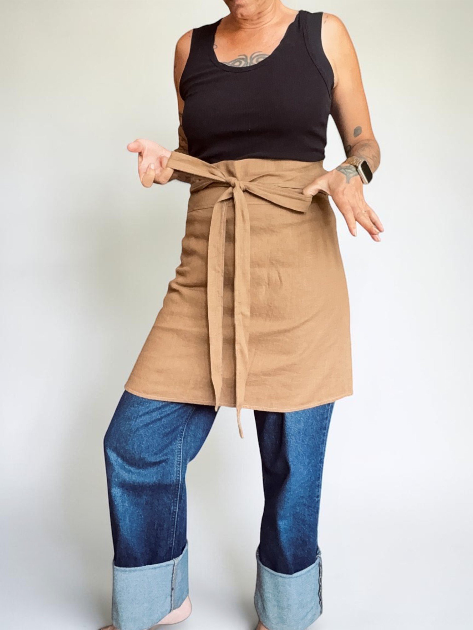 A woman wearing a black sleeveless top and a ginger linen amphitrite cafe apron standing against a plain white wall.