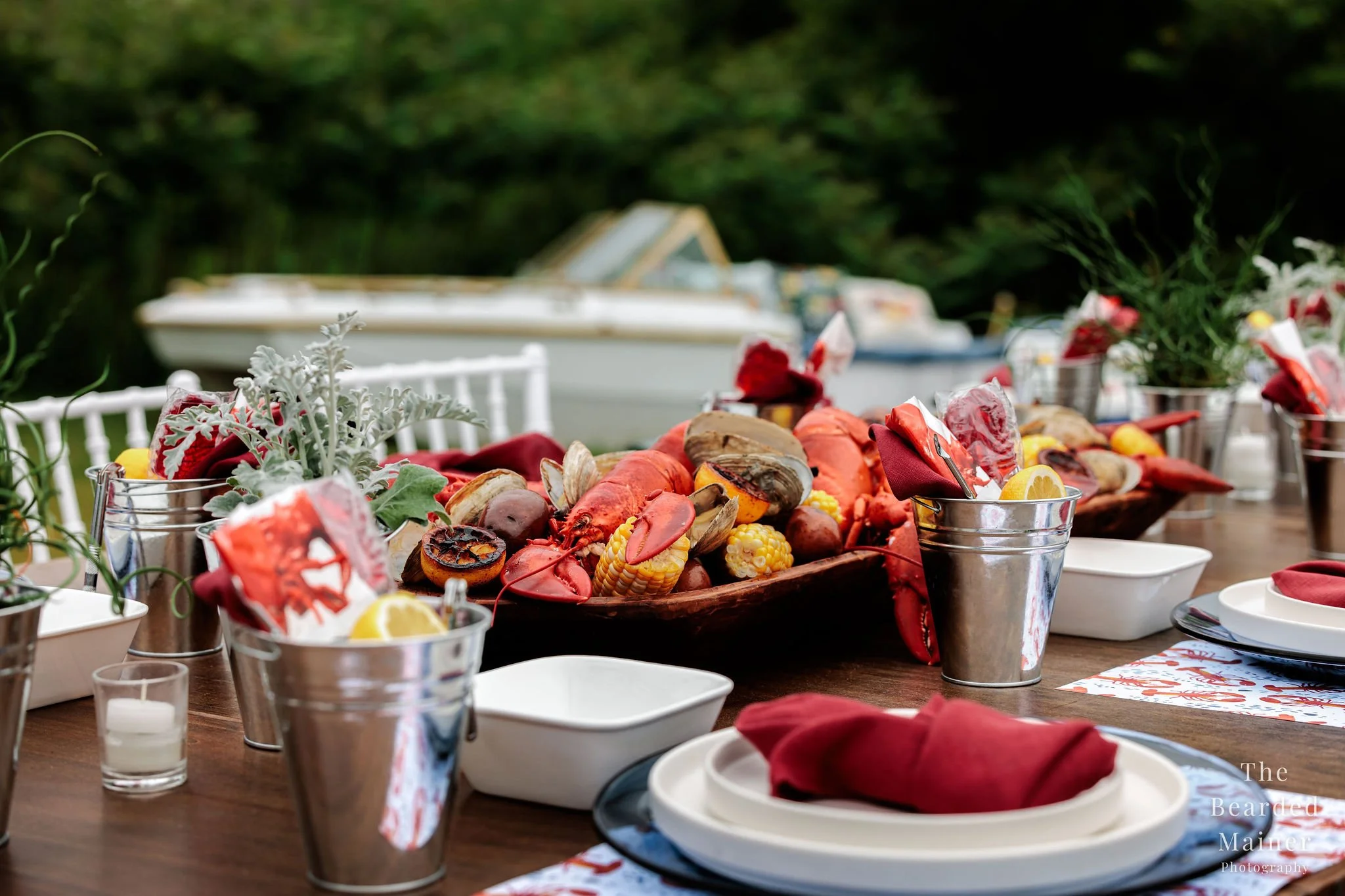 Outdoor table set with lobster, corn, and seafood, decorated with red amphitrite linen napkins, small potted plants, and white plates, with a blurred boat and greenery in the background.