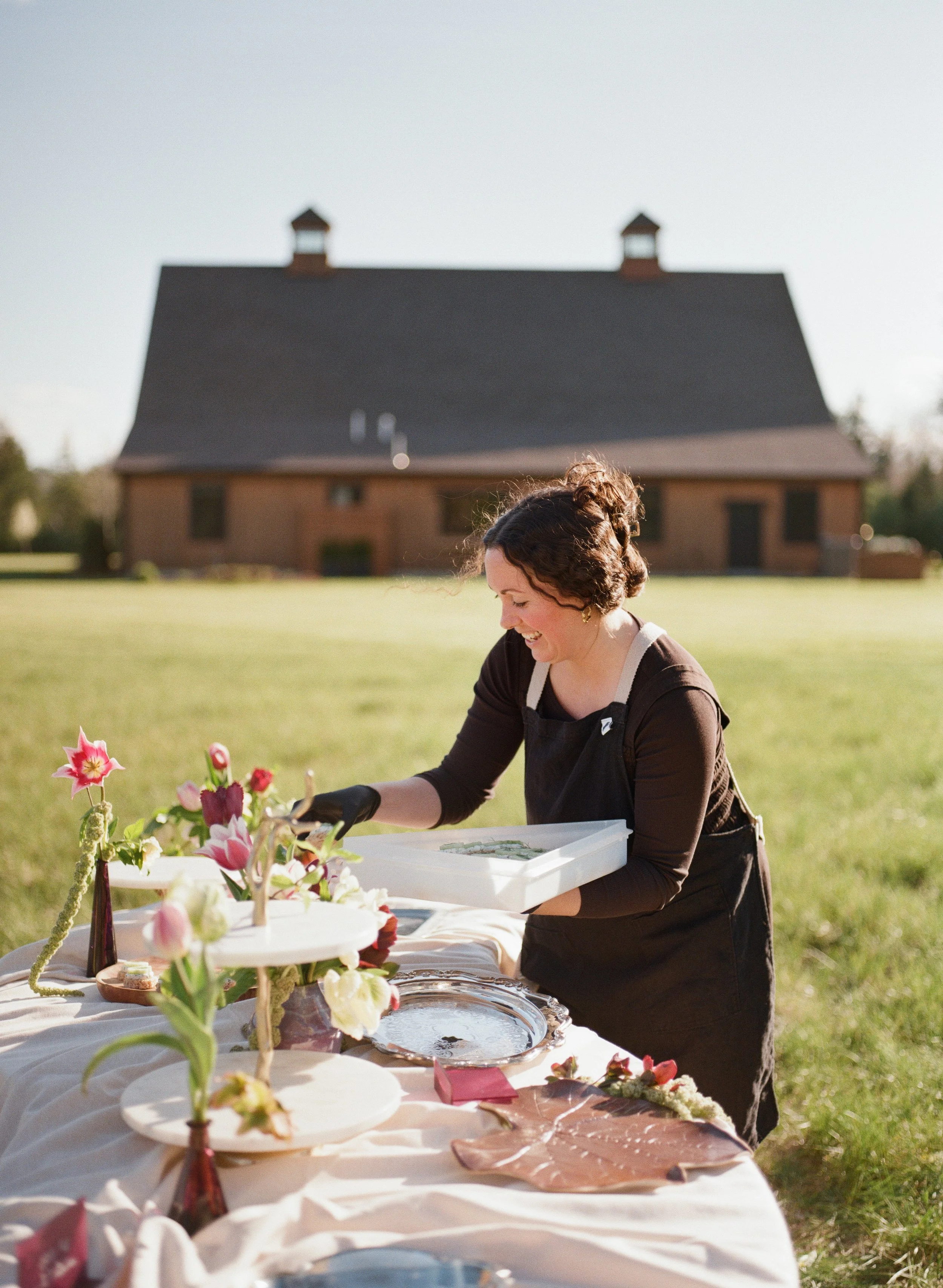 A chef in a black amphitrite apron arranging colorful flowers at an outdoor table with a variety of flower arrangements and desserts, with a large house in the background on a sunny day.