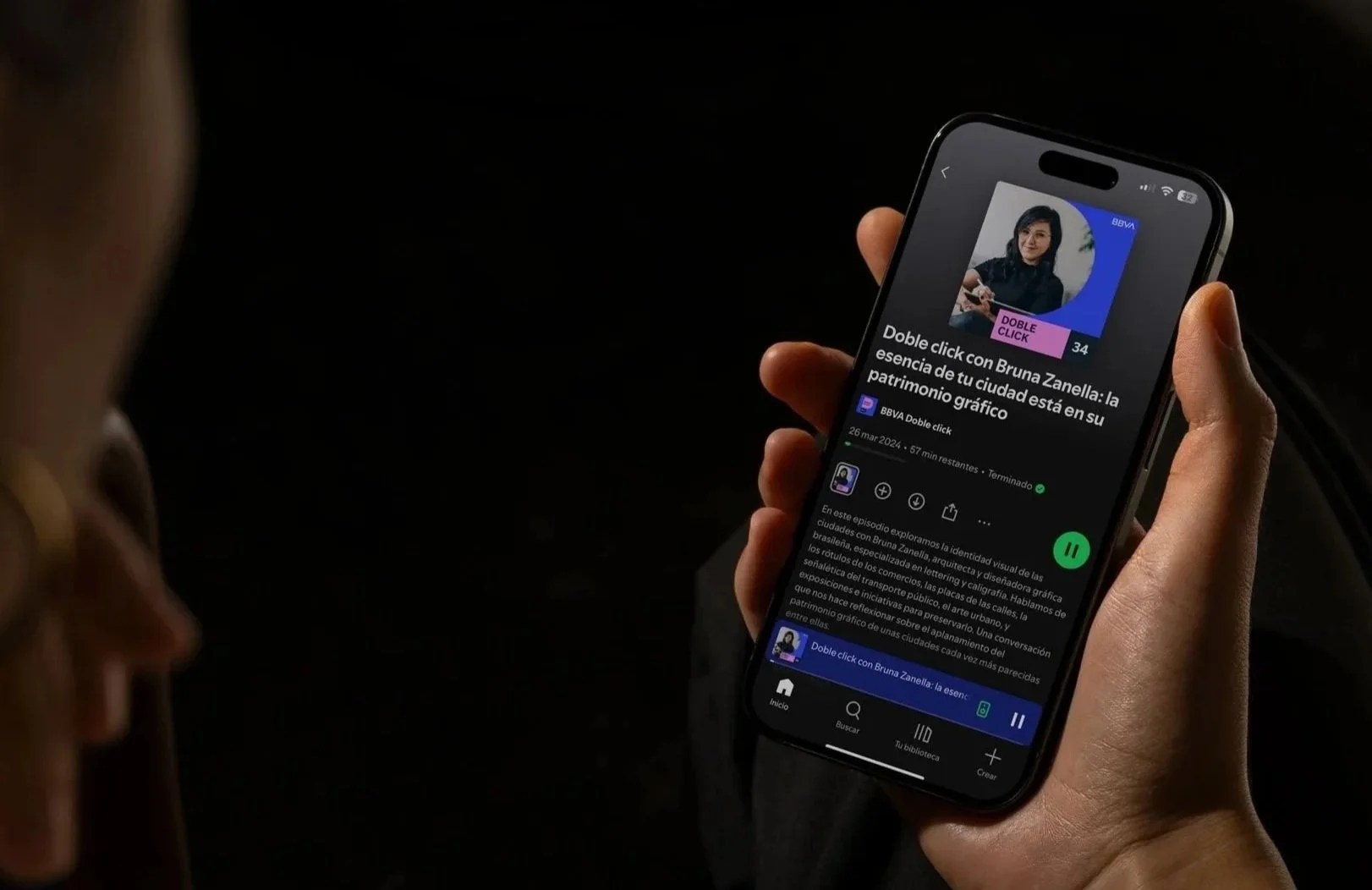 Persona sosteniendo un teléfono móvil que muestra un podcast titulado 'Doble click con Bruna Zanella' en una pantalla con fondo negro.