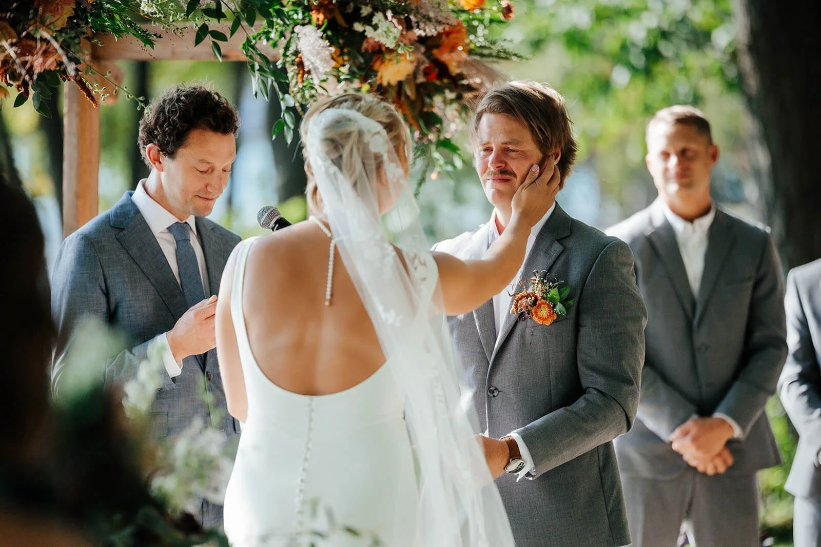 White Birch Lodge wedding photographer Elk Rapids Michigan bride wiping groom's tears during outdoor ceremony under floral arch, top Northern Michigan documentary wedding photography