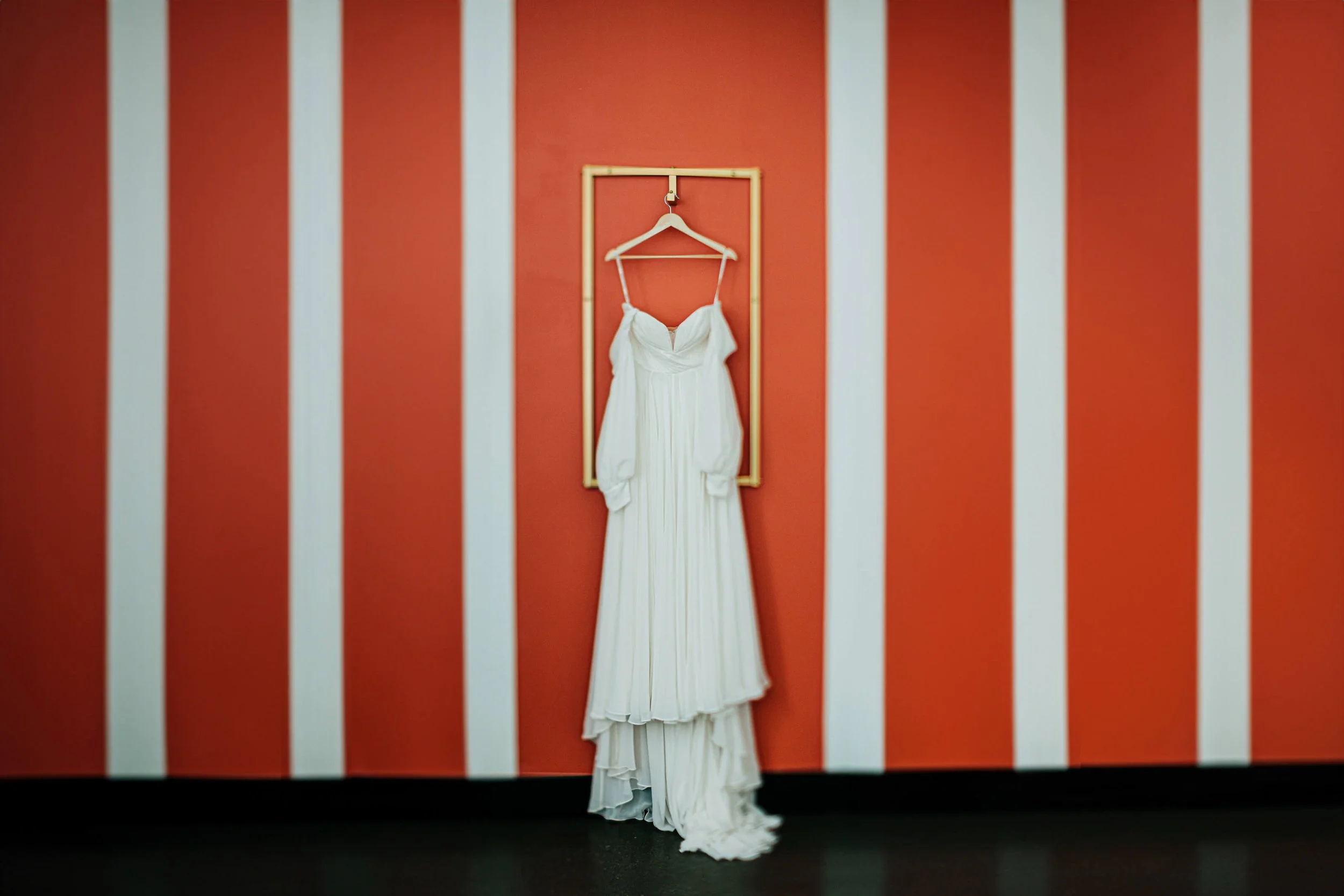 Leona Road wedding photographer Grand Rapids Michigan editorial white bridal gown hanging on gold rack against bold red and white striped wall, best Grand Rapids wedding photography