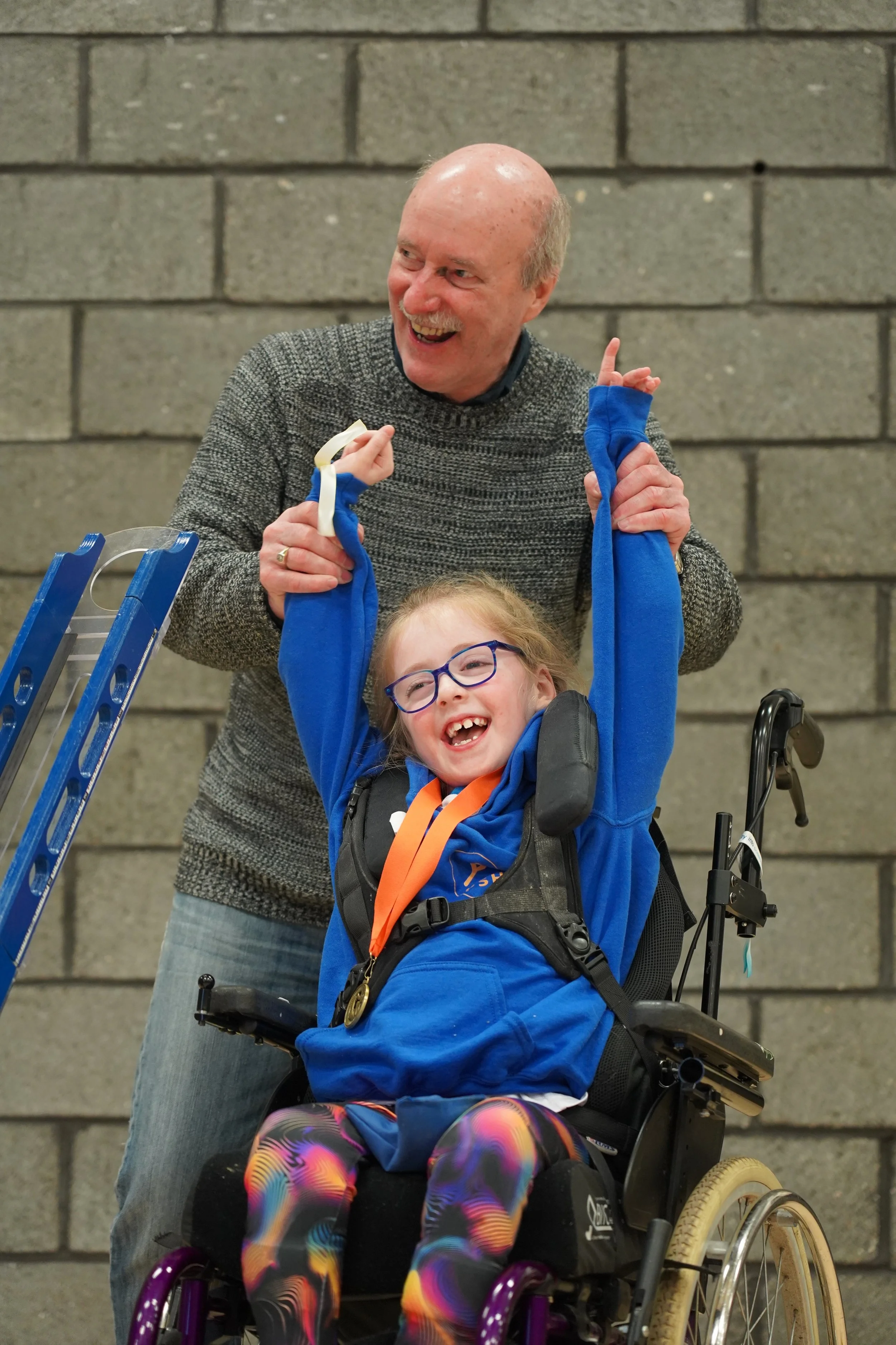 Young girl and dad playing boccia. The dad is assisting the young girl. She is in a wheelchair and is using a boccia ramp. Both are smiling and the dad is raising her arms in the air in triumph.