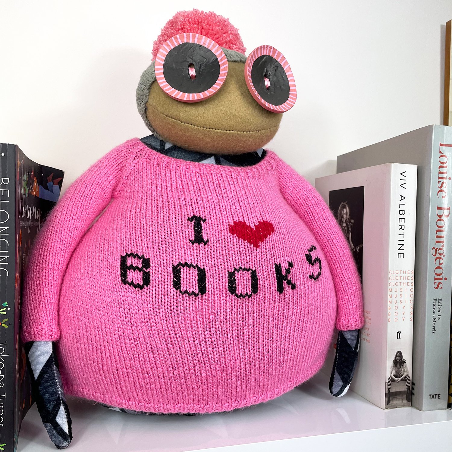 Handmade Ruby Ruth Doll named Burt wearing a bright pink jumper with embroidered text that says ‘I heart books’, sitting between books.