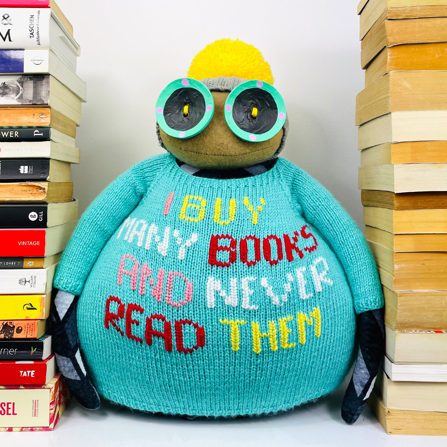 Limited edition Ruby Ruth Doll named Burt wearing a turquoise jumper with embroidered text that reads 'I buy many books and never read them', surrounded by stacks of books.
