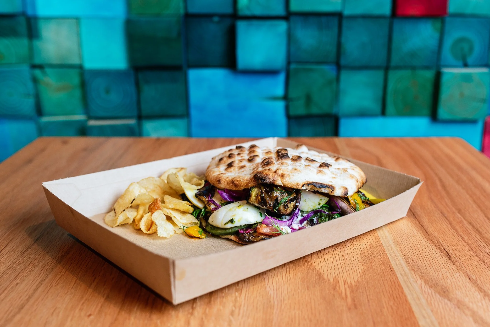 A food tray with a sandwich and potato chips on a wooden table in front of a colorful, abstract background.