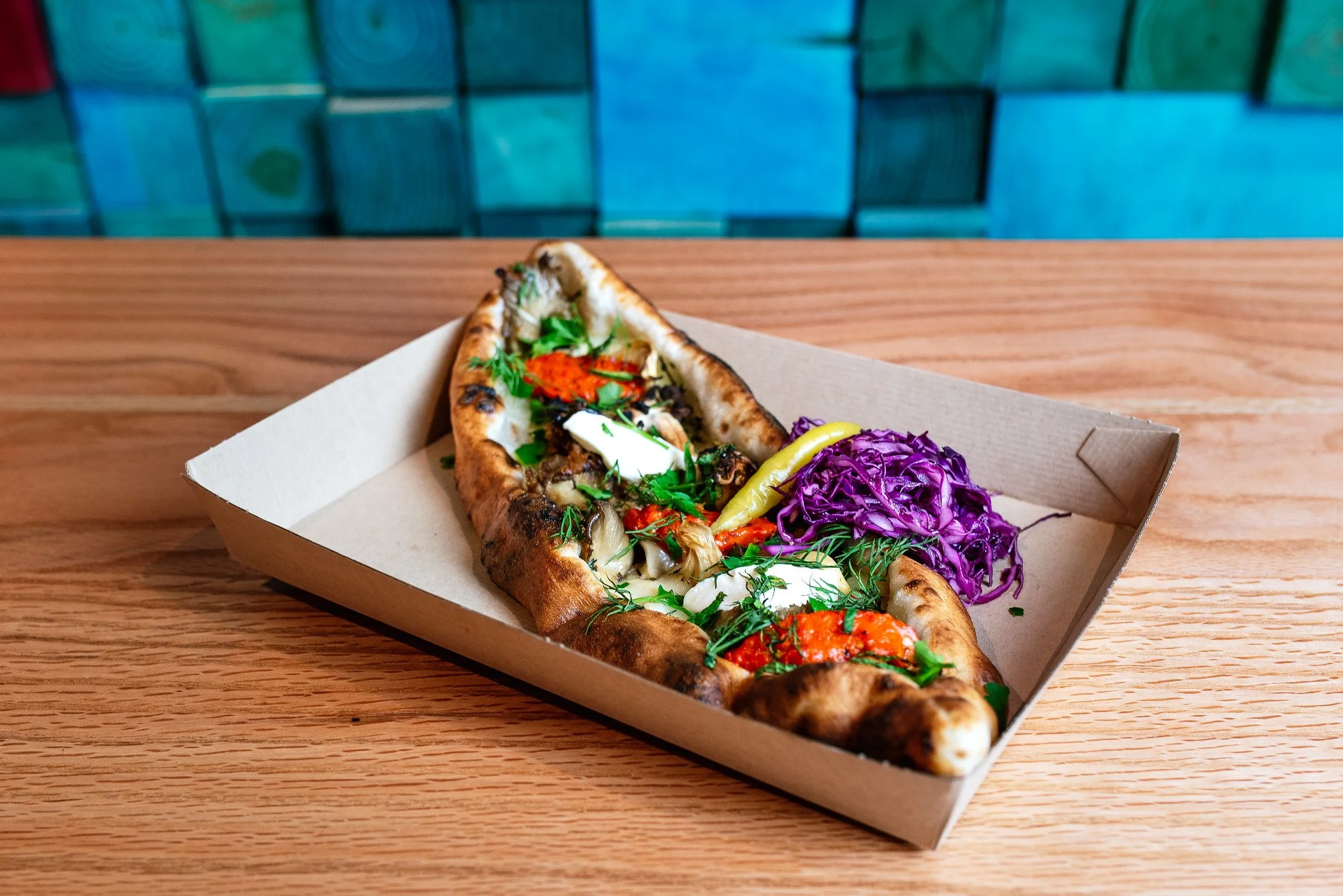 Veggie pizza with purple cabbage slaw and pickled peppers in a cardboard tray on a wooden table.
