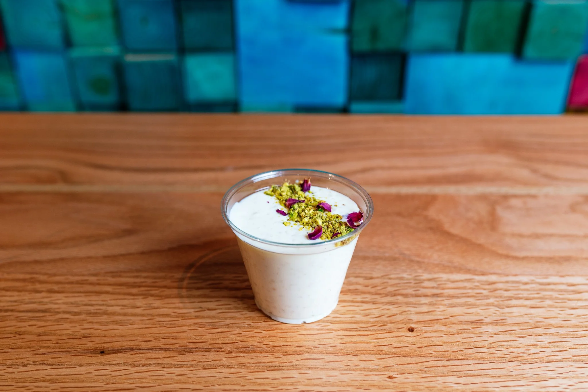A small transparent plastic cup filled with a creamy white dessert, garnished with crushed pistachios and rose petals, placed on a wooden table with a colorful tiled wall in the background.